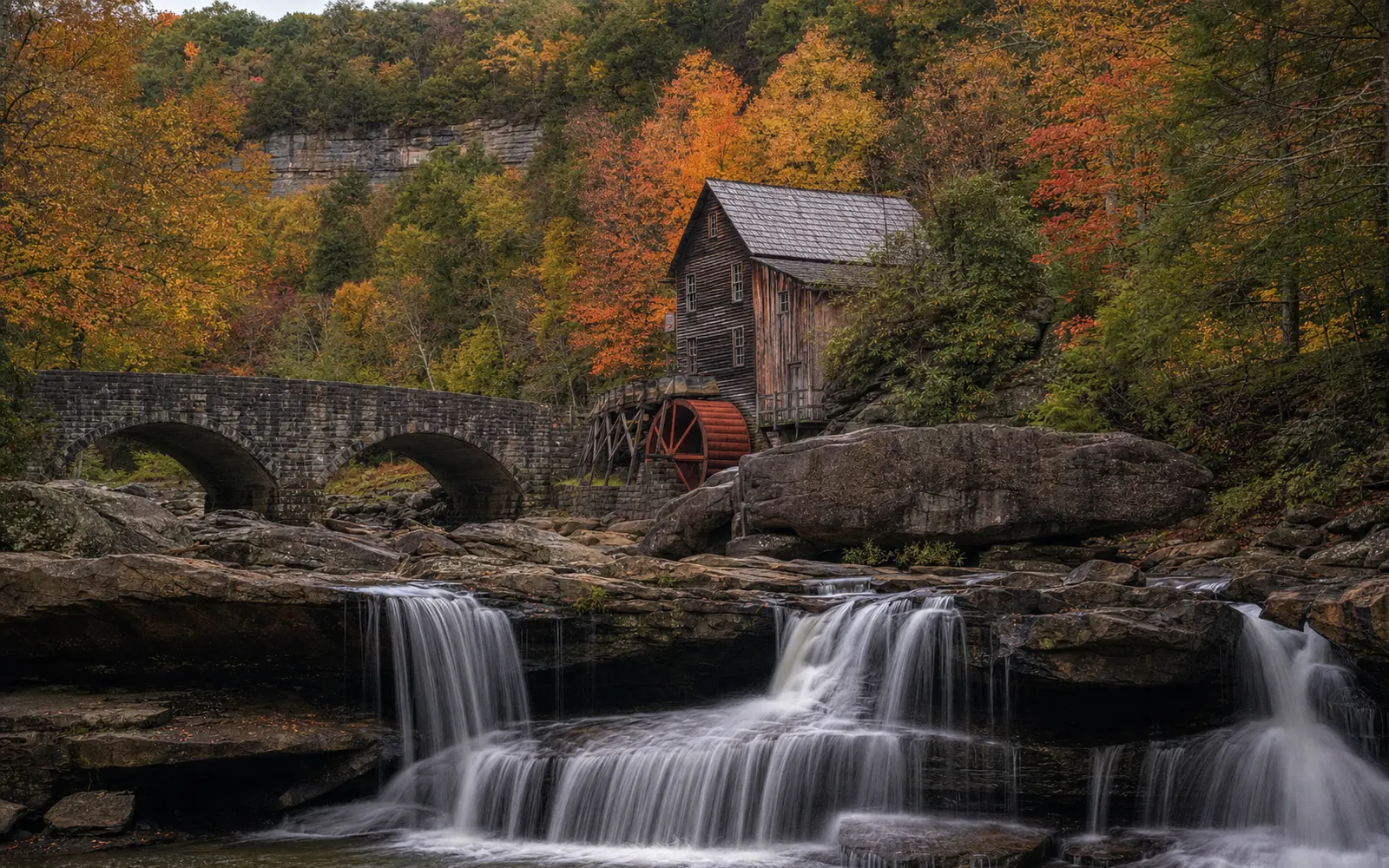 The weathered wooden Glade Creek Grist Mill with its red water wheel beside a boulder-filled creek and a stone footbridge, AI-generated illustration