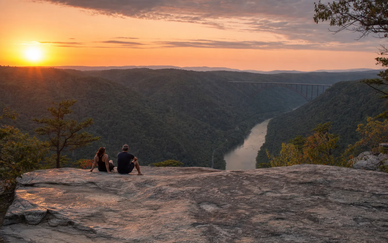 A bare sandstone ledge on the gorge rim at sunset, two adults seated back from the edge, the river below and the bridge a small arch in the distance, AI-generated illustration