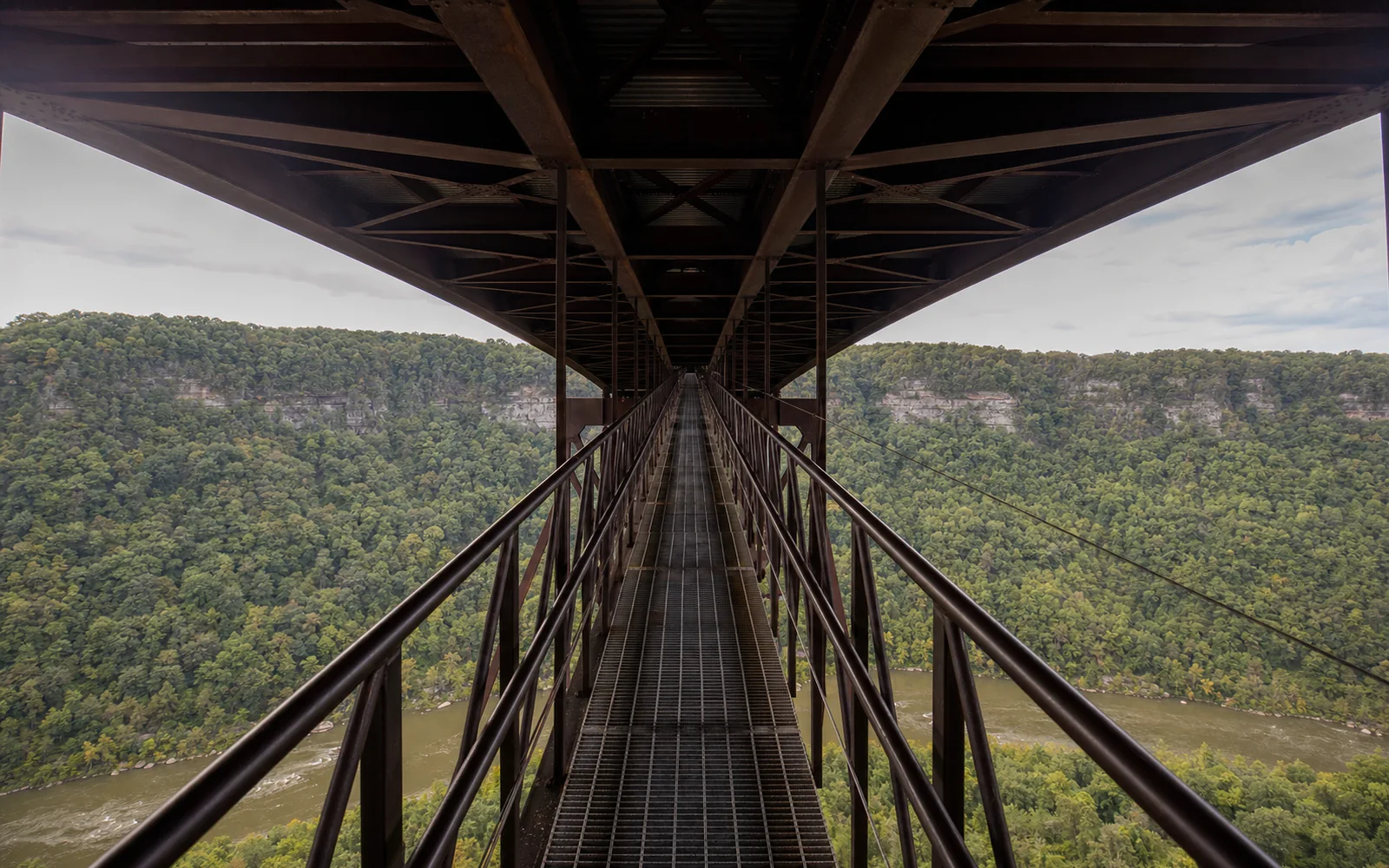The Bridge Walk catwalk under the New River Gorge Bridge deck in long perspective, a narrow grated steel walkway beneath steel stringers and cross-bracing with the gorge beyond the railing, AI-generated illustration