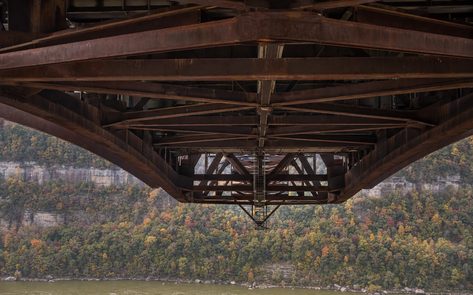 The underside of the New River Gorge Bridge's deck seen from the Canyon Rim lower overlook with a long lens, the thin catwalk running beneath the deck trusses across the span, AI-generated illustration