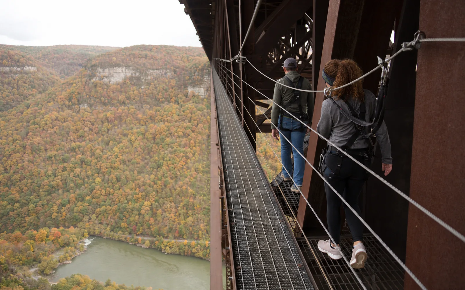 Two harnessed walkers on the narrow steel catwalk under the deck of the New River Gorge Bridge, a safety cable overhead and the river far below, AI-generated illustration