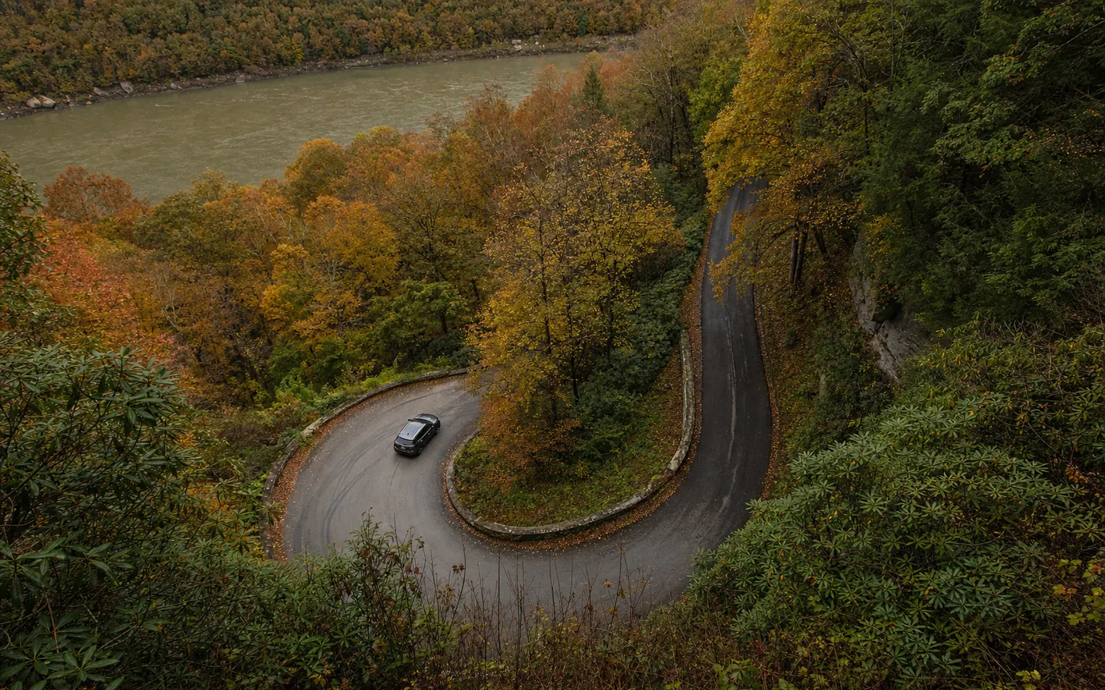 A tight hairpin switchback on narrow one-lane Fayette Station Road descending through hemlock and rhododendron, a stone retaining wall, one car, AI-generated illustration