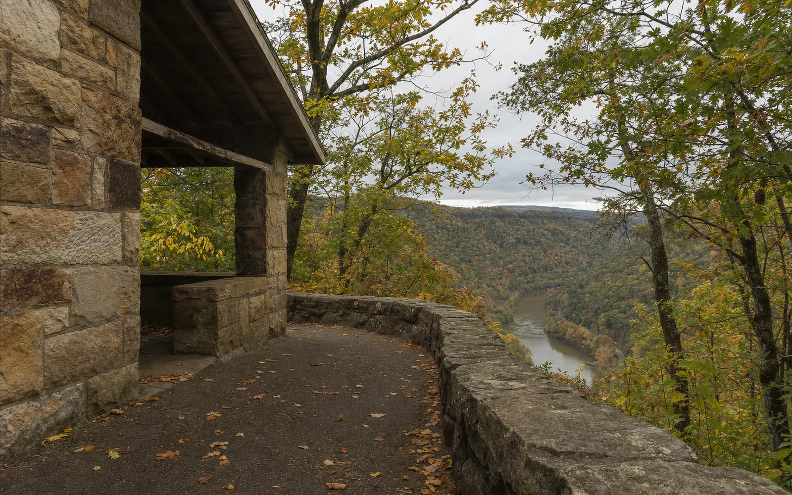 The Civilian Conservation Corps stone overlook shelter at Hawks Nest State Park, rough-cut sandstone walls, a low stone parapet at the cliff edge, the New River's bend visible far below through the trees, AI-generated illustration