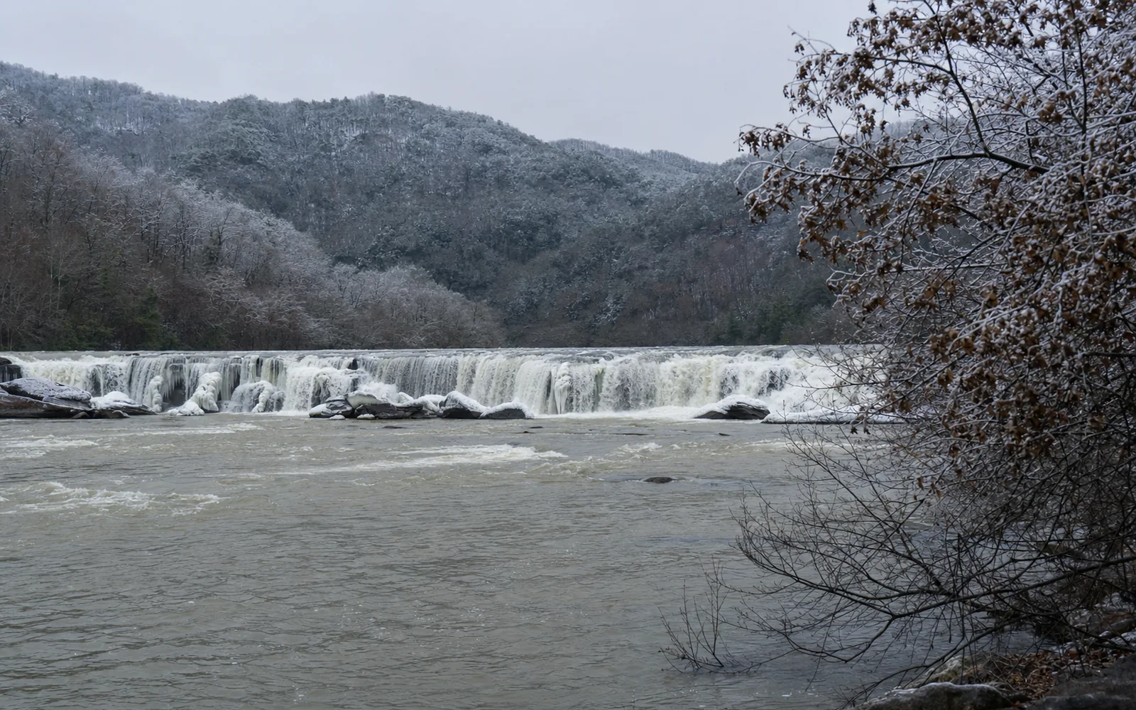 Sandstone Falls in winter, the river-wide low ledges with ice along their lips, water pouring between frosted wooded islands, grey sky, AI-generated illustration
