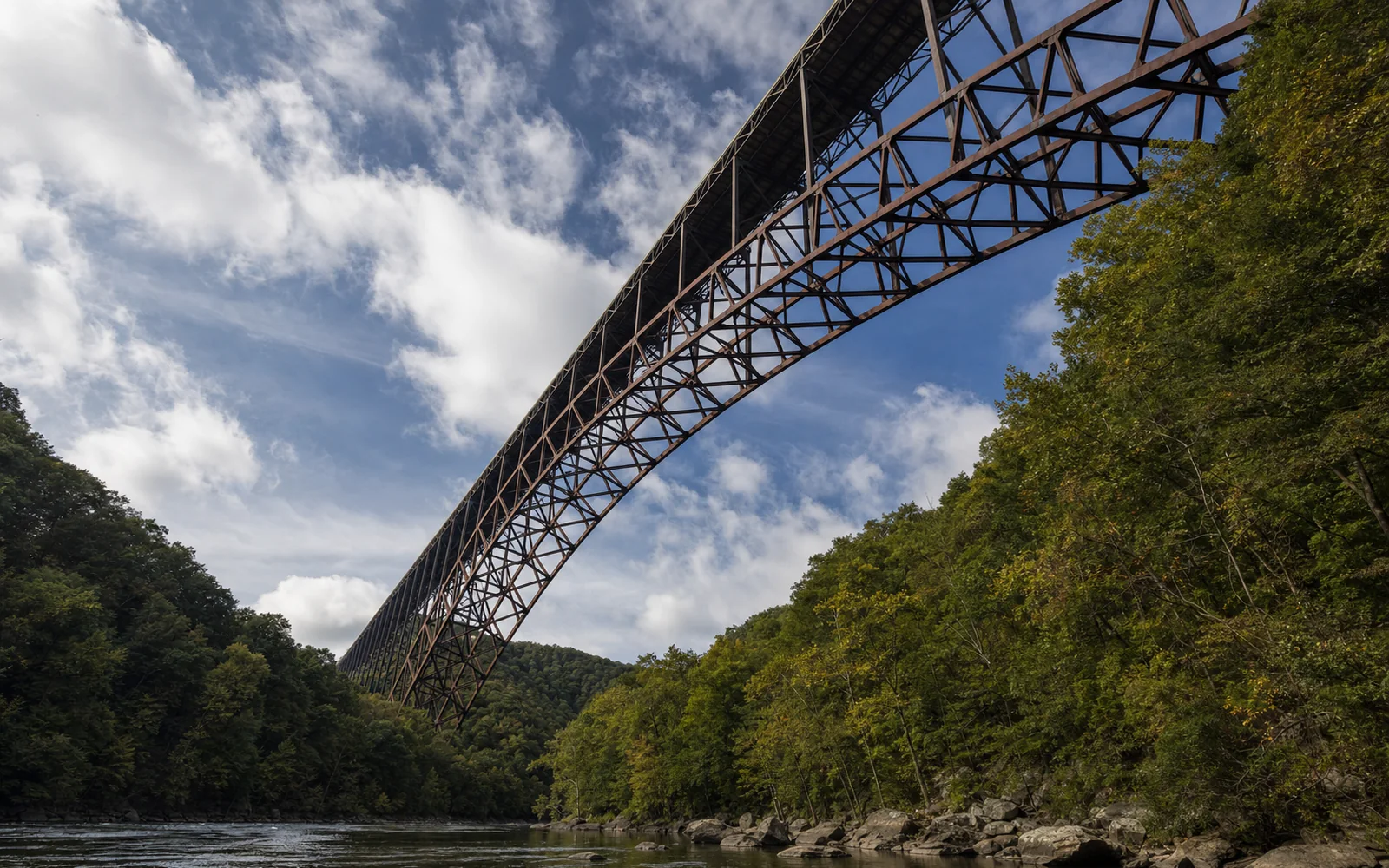 The rust-brown steel arch and deck trusses of the New River Gorge Bridge seen from below on Fayette Station Road, with forest on both slopes of the gorge, AI-generated illustration