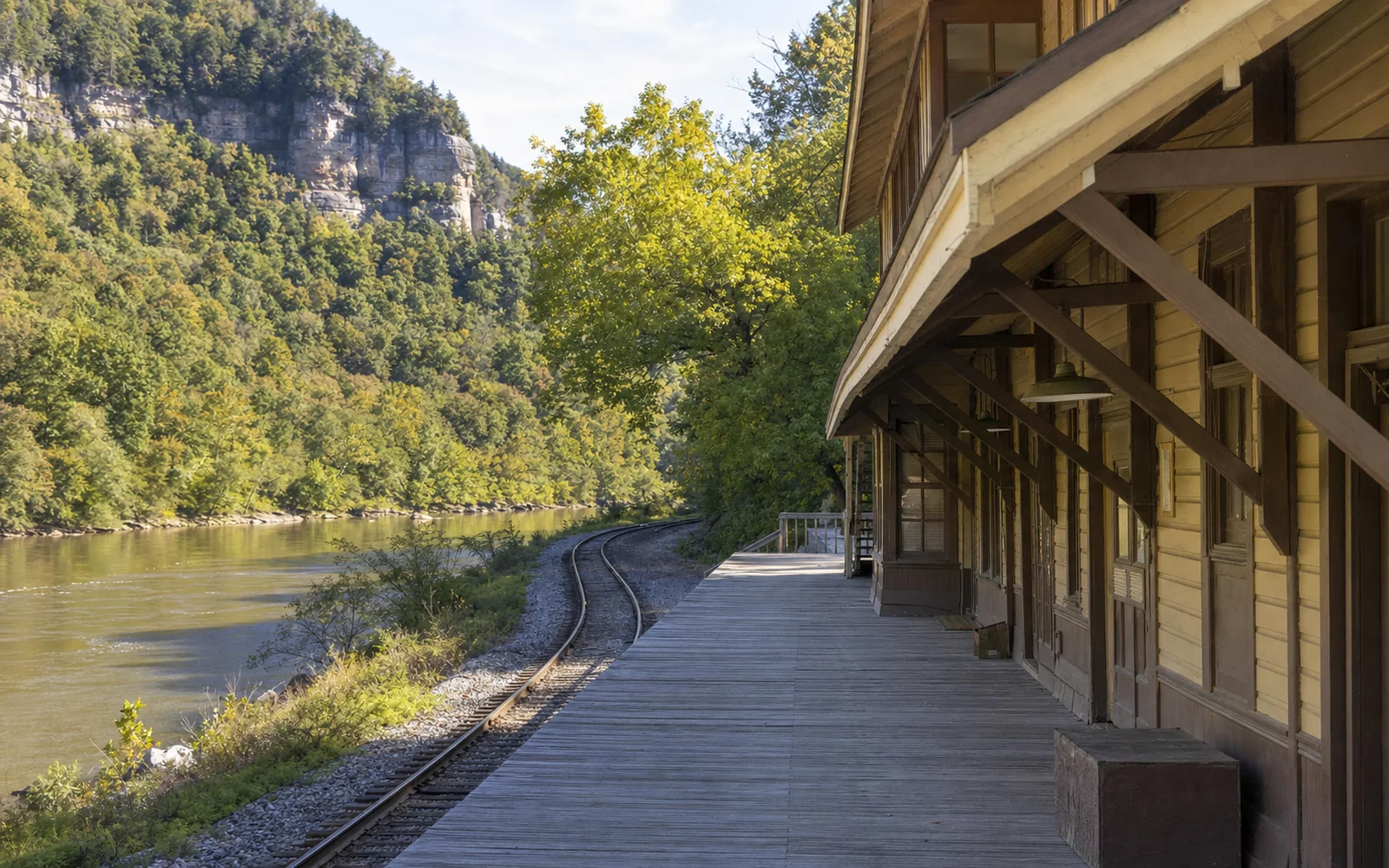 The empty platform of the restored Thurmond depot, a wooden two-story station with a deep roof overhang on brackets, the railroad tracks curving along the river, AI-generated illustration