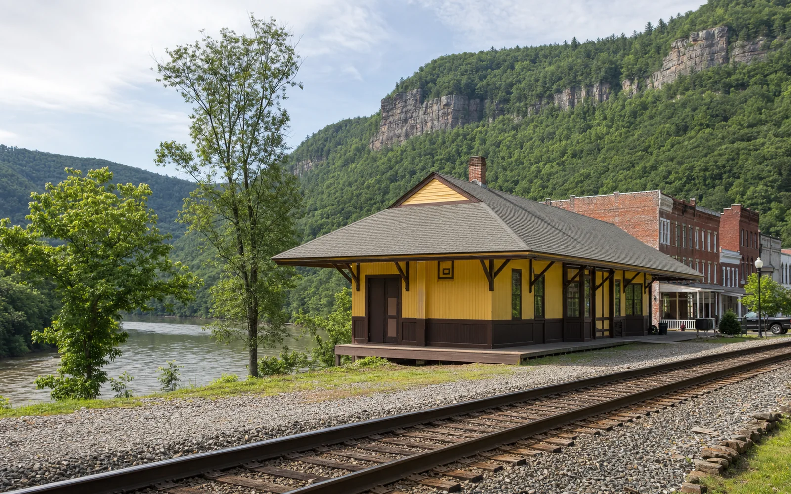 The restored Thurmond depot beside the railroad tracks, with a row of brick commercial buildings, the river and wooded cliffs behind, AI-generated illustration
