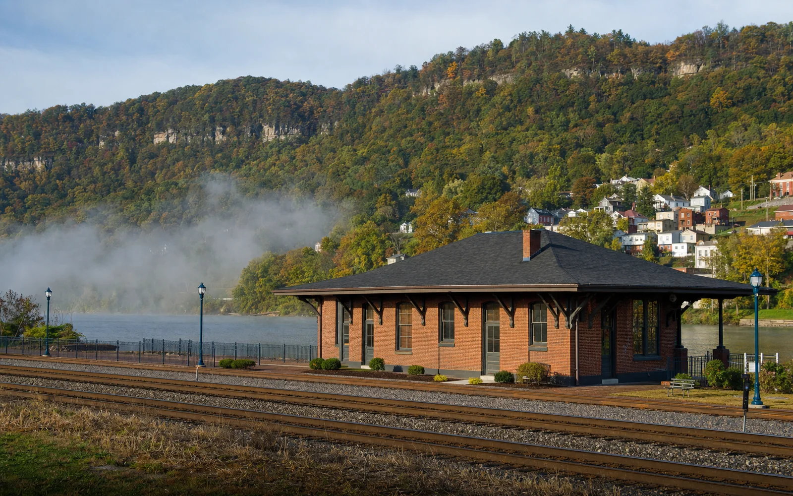 A single-story brick railroad depot beside the tracks at Hinton with the wide New River behind and houses climbing the hillside, AI-generated illustration