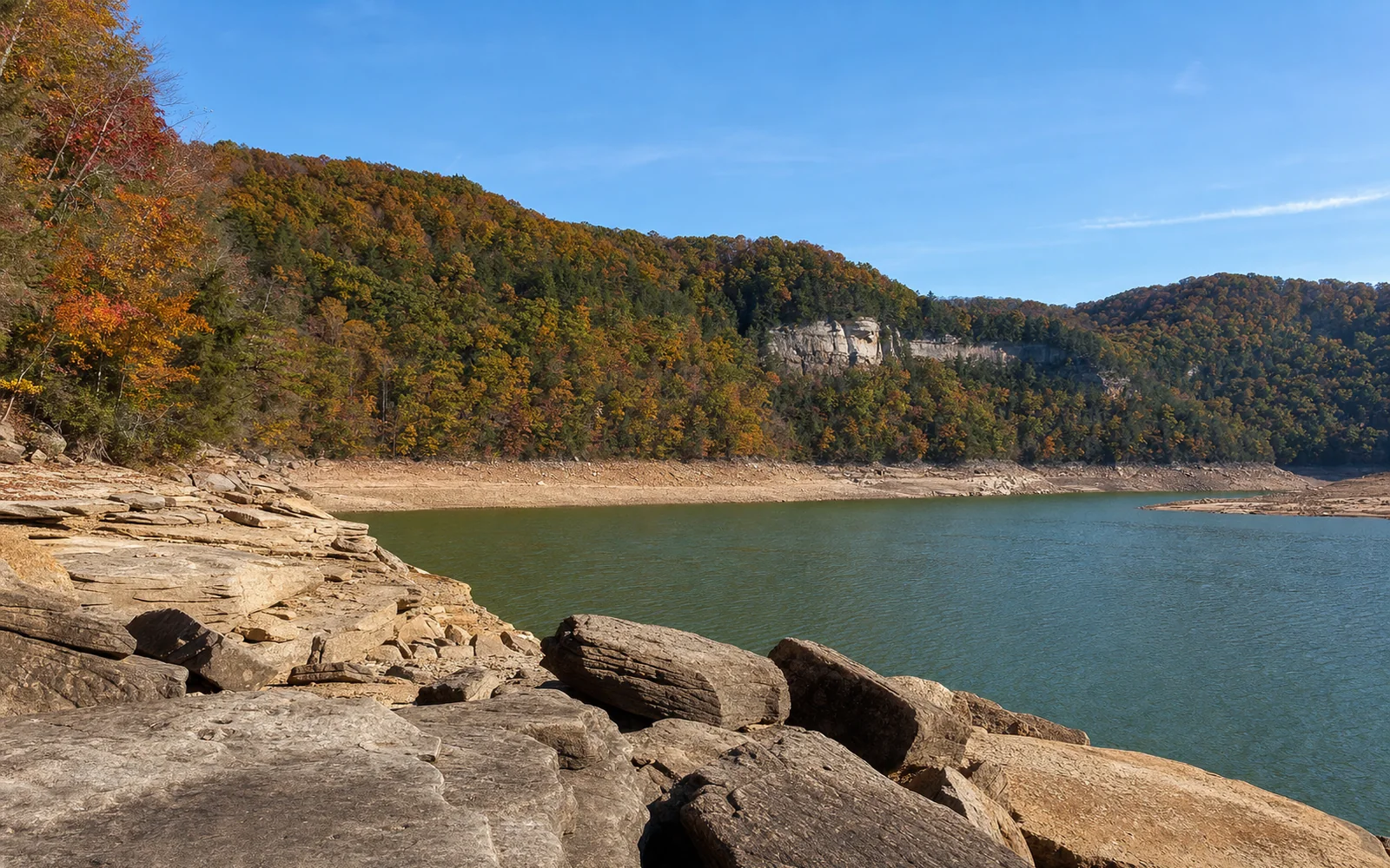 Summersville Lake at the autumn drawdown, exposed pale sandstone ledges and boulders along the shoreline, the lower green water, forested hills, AI-generated illustration
