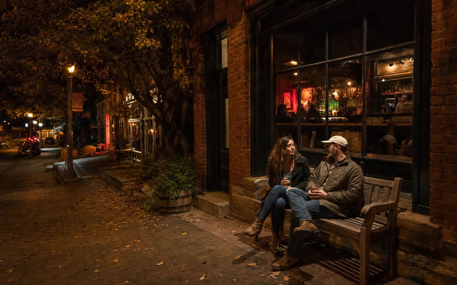 A small-town brick street at night with a warmly lit bar window and two adults on a bench outside, AI-generated illustration