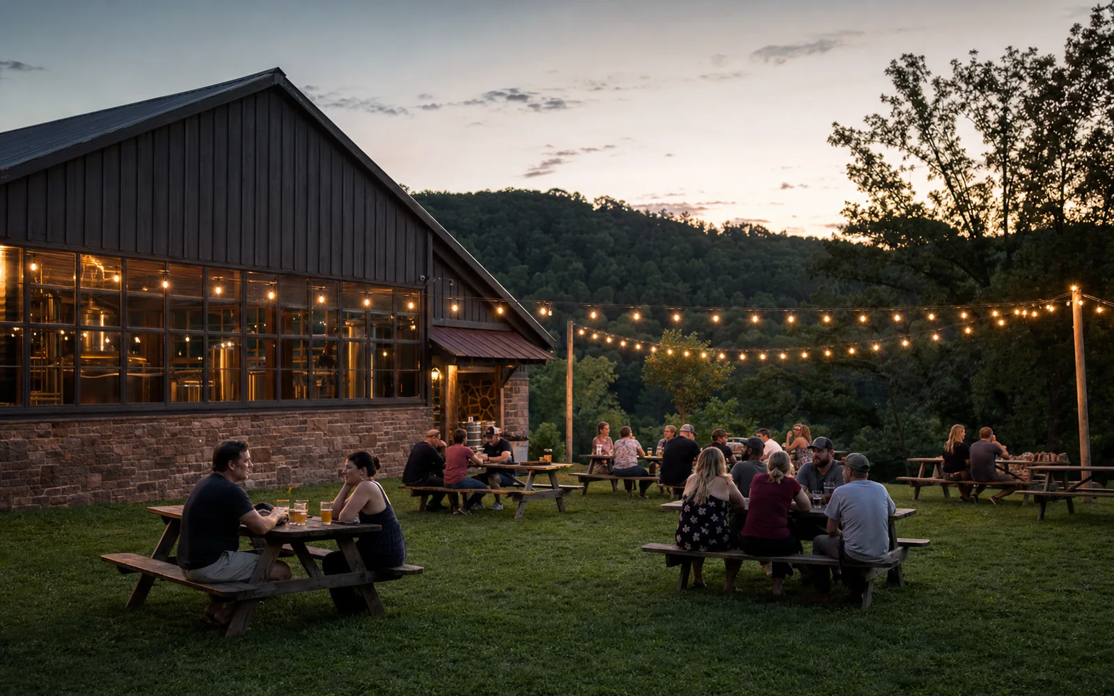 Adults at picnic tables on a grassy lawn outside a rural brewery barn at dusk, string lights, wooded hillside, AI-generated illustration