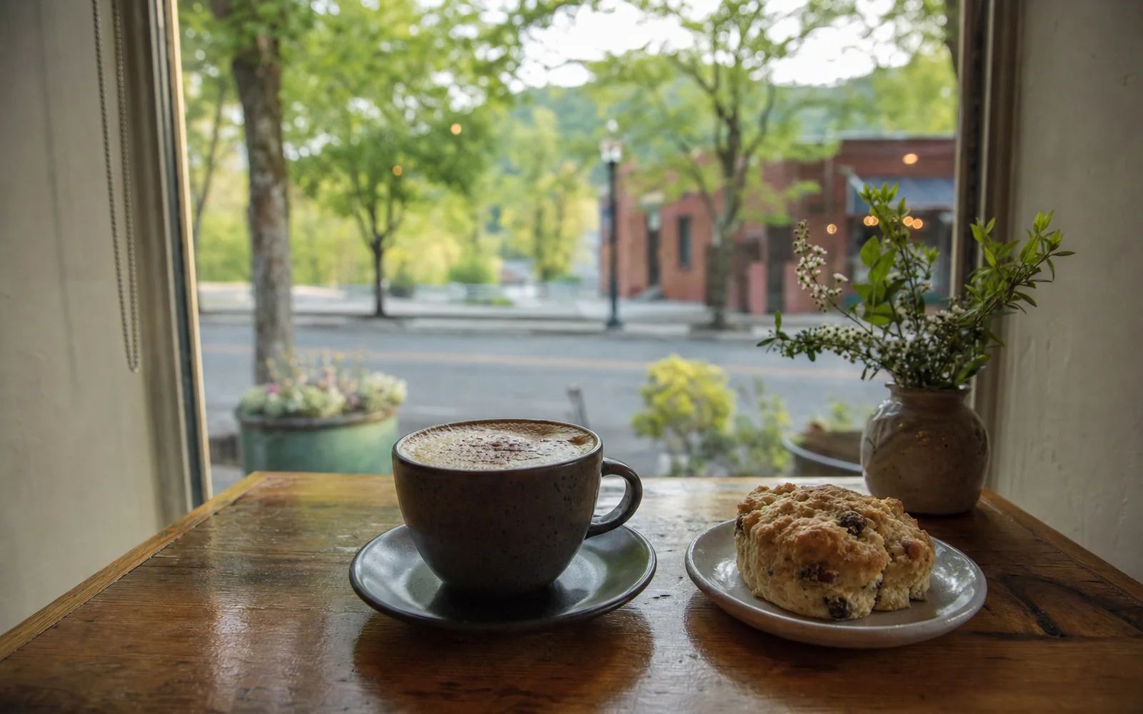 A cappuccino and a scone on a small cafe table by a window looking out on a leafy small-town street, AI-generated illustration
