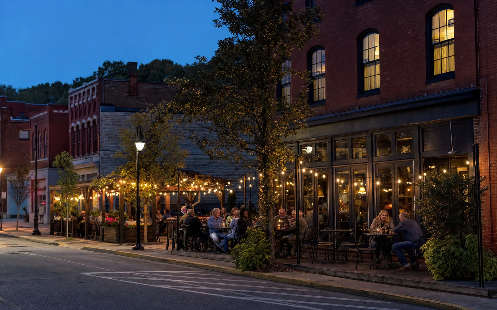 Adults dining at string-lit patio tables outside red brick storefronts on a small-town main street at dusk, with wooded hills behind, AI-generated illustration
