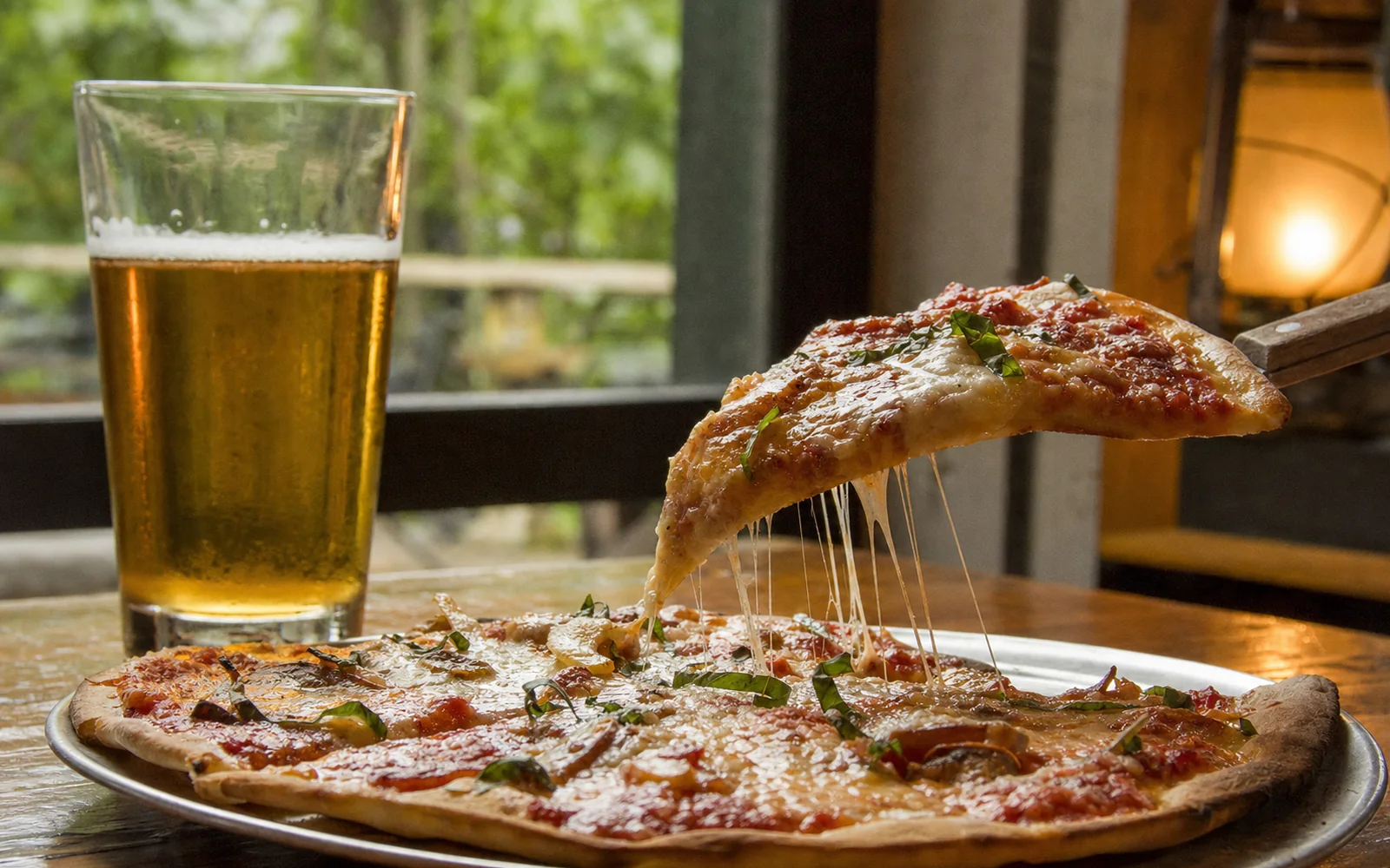 A slice of thin-crust pizza lifted from a round pie with a stretch of cheese, a pint of lager beside it on a wooden table, AI-generated illustration