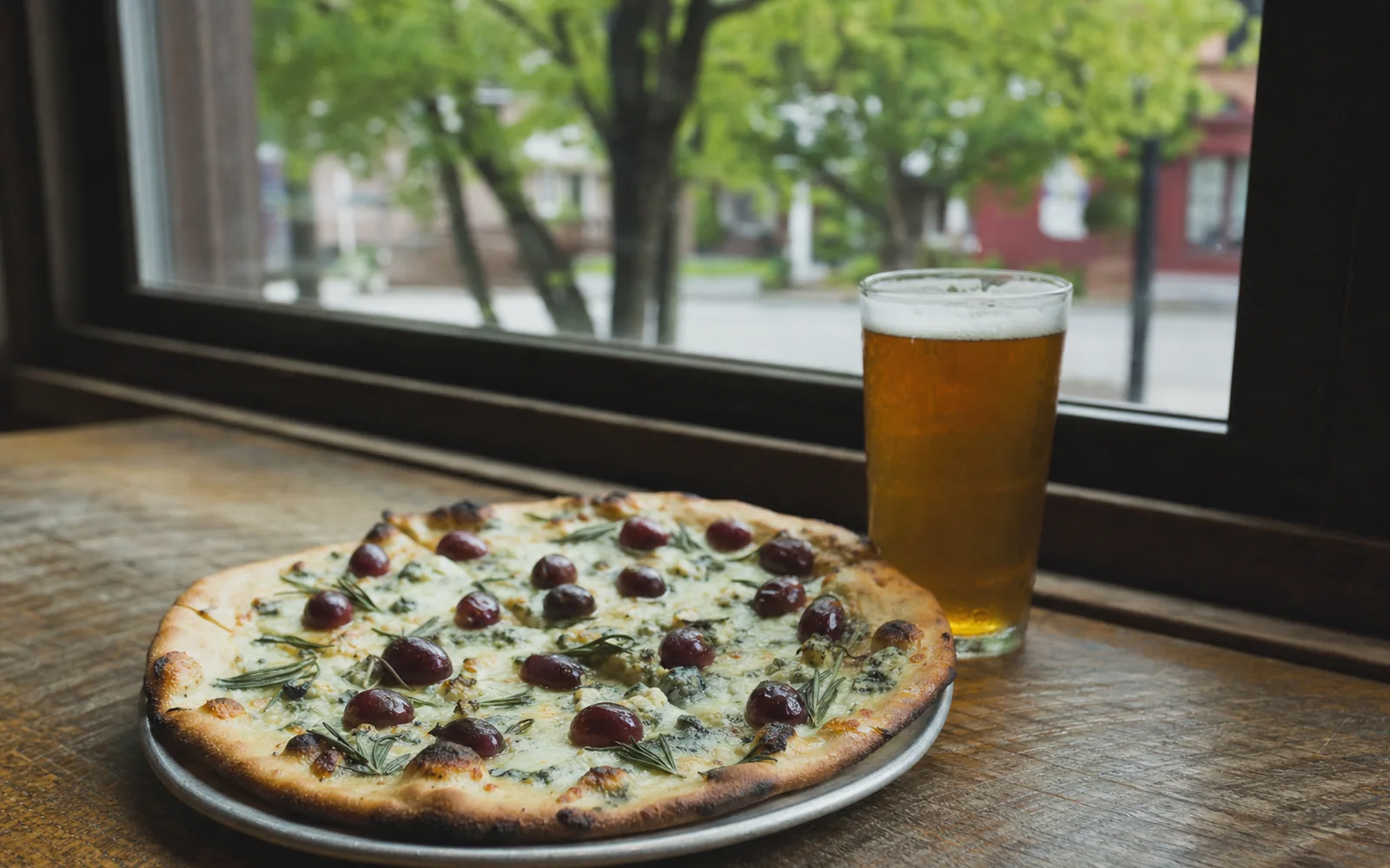 A thin-crust pizza topped with red grapes and crumbled blue cheese on a table by a window that looks onto a small-town street of brick buildings and trees, AI-generated illustration