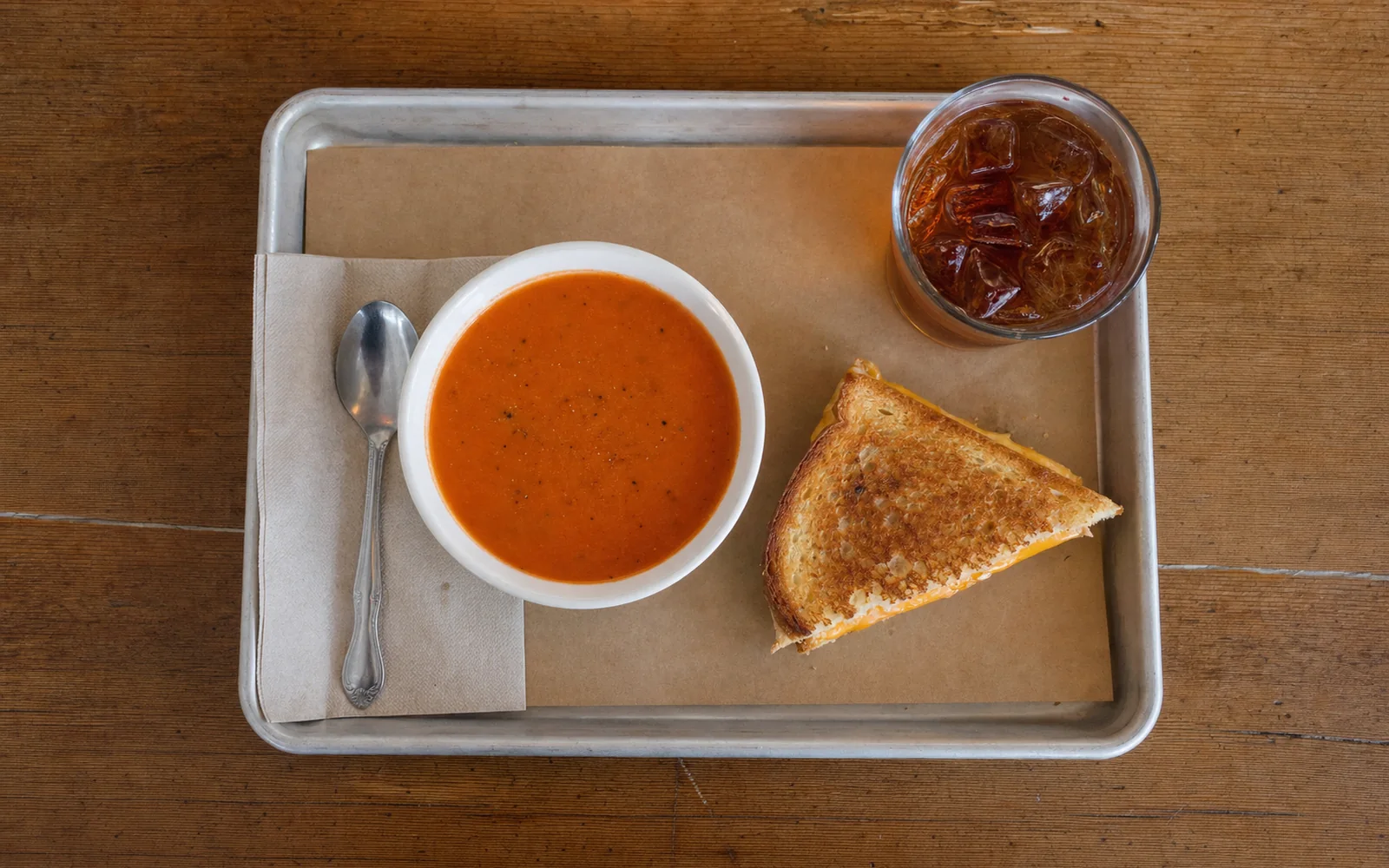 A bowl of tomato soup and half a grilled sandwich on a tray with an iced tea, AI-generated illustration