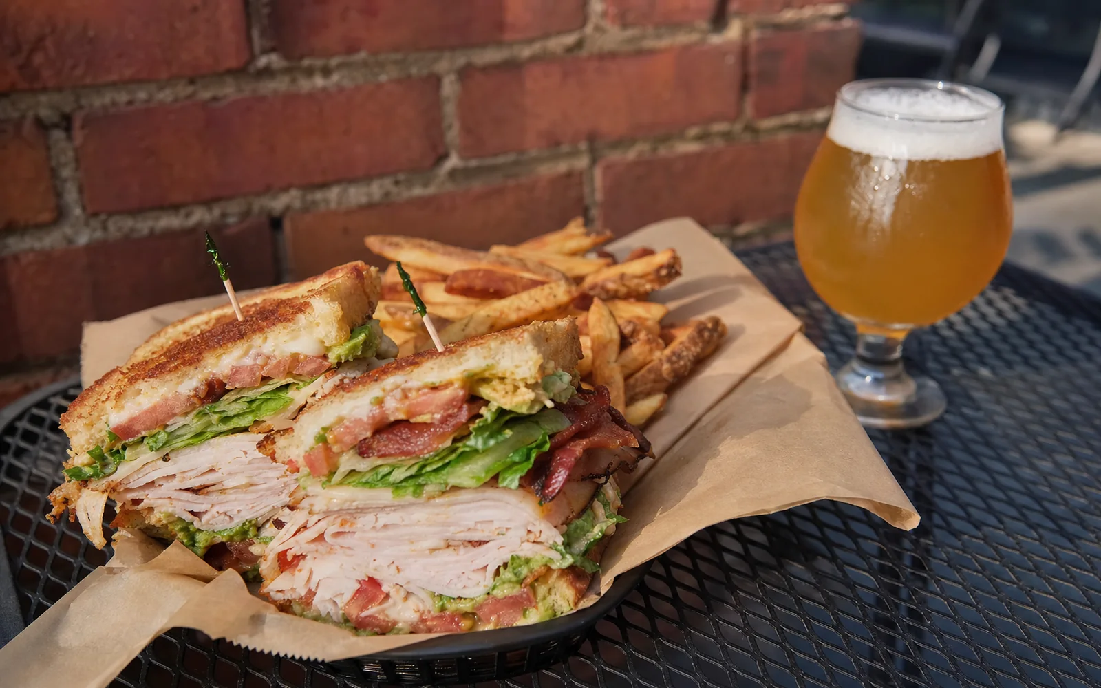 A stacked club sandwich and a glass of amber beer on a patio table against a red brick wall, AI-generated illustration