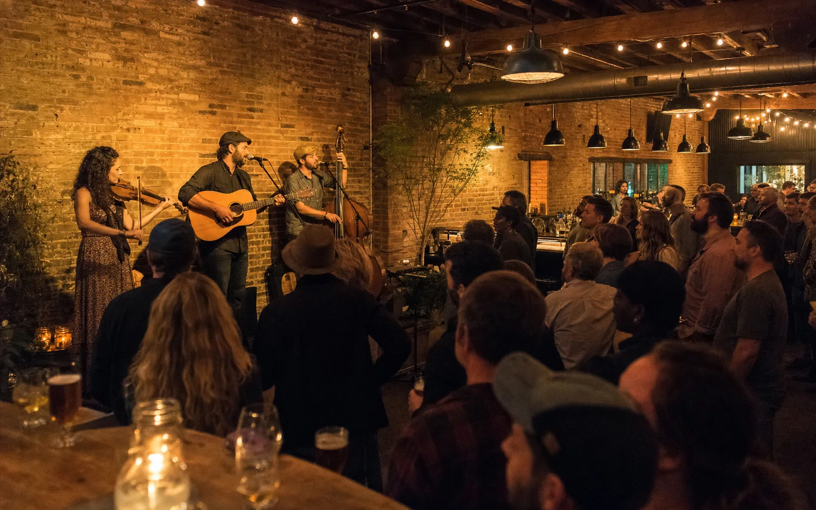 A fiddle, guitar and upright bass trio plays against a brick wall to a standing crowd in a dim bar room hung with string lights, AI-generated illustration