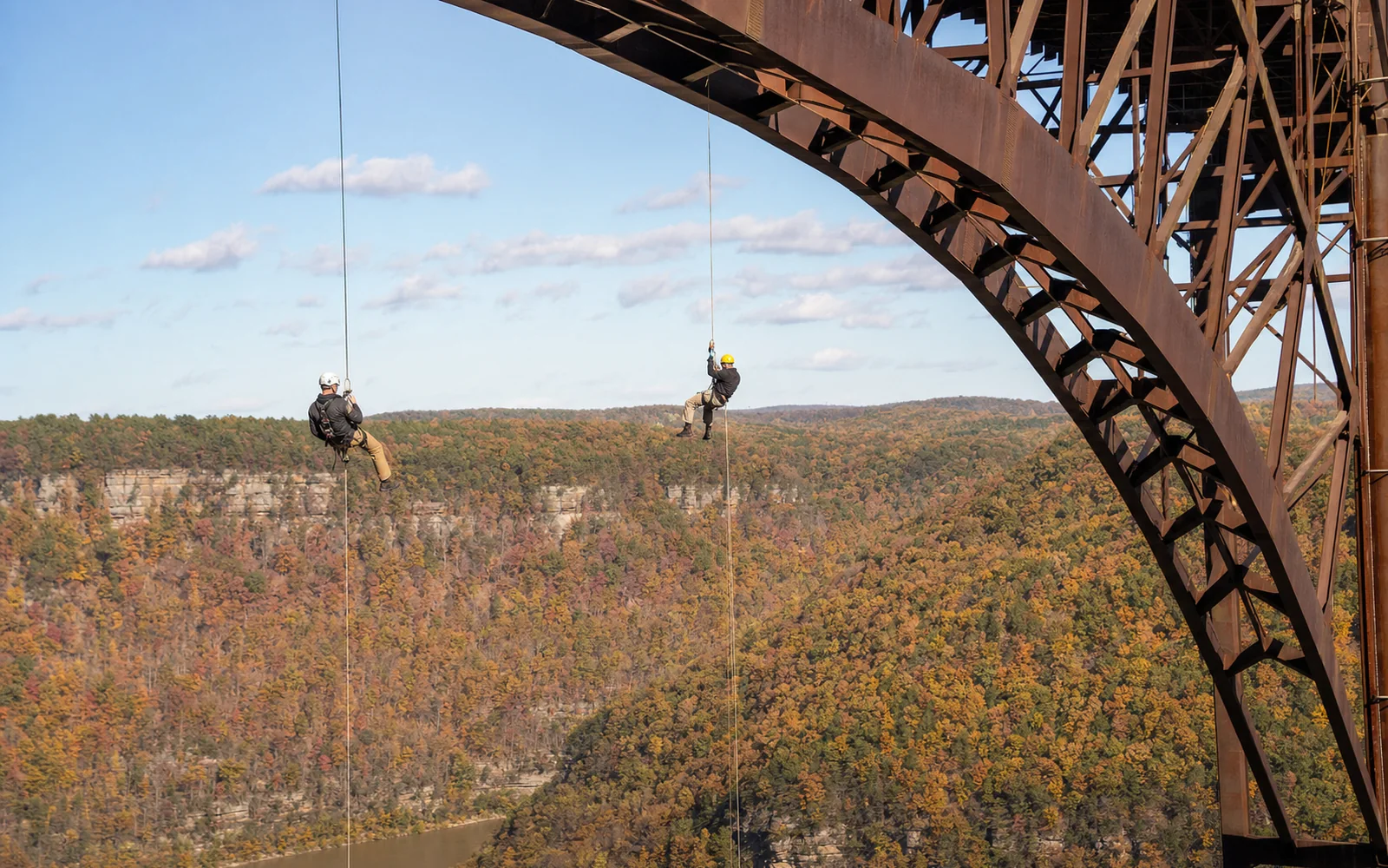 Bridge Day rappellers, two adult climbers in helmets descending long ropes hung beneath the New River Gorge Bridge's deck, the steel arch overhead, the gorge in fall color below, AI-generated illustration