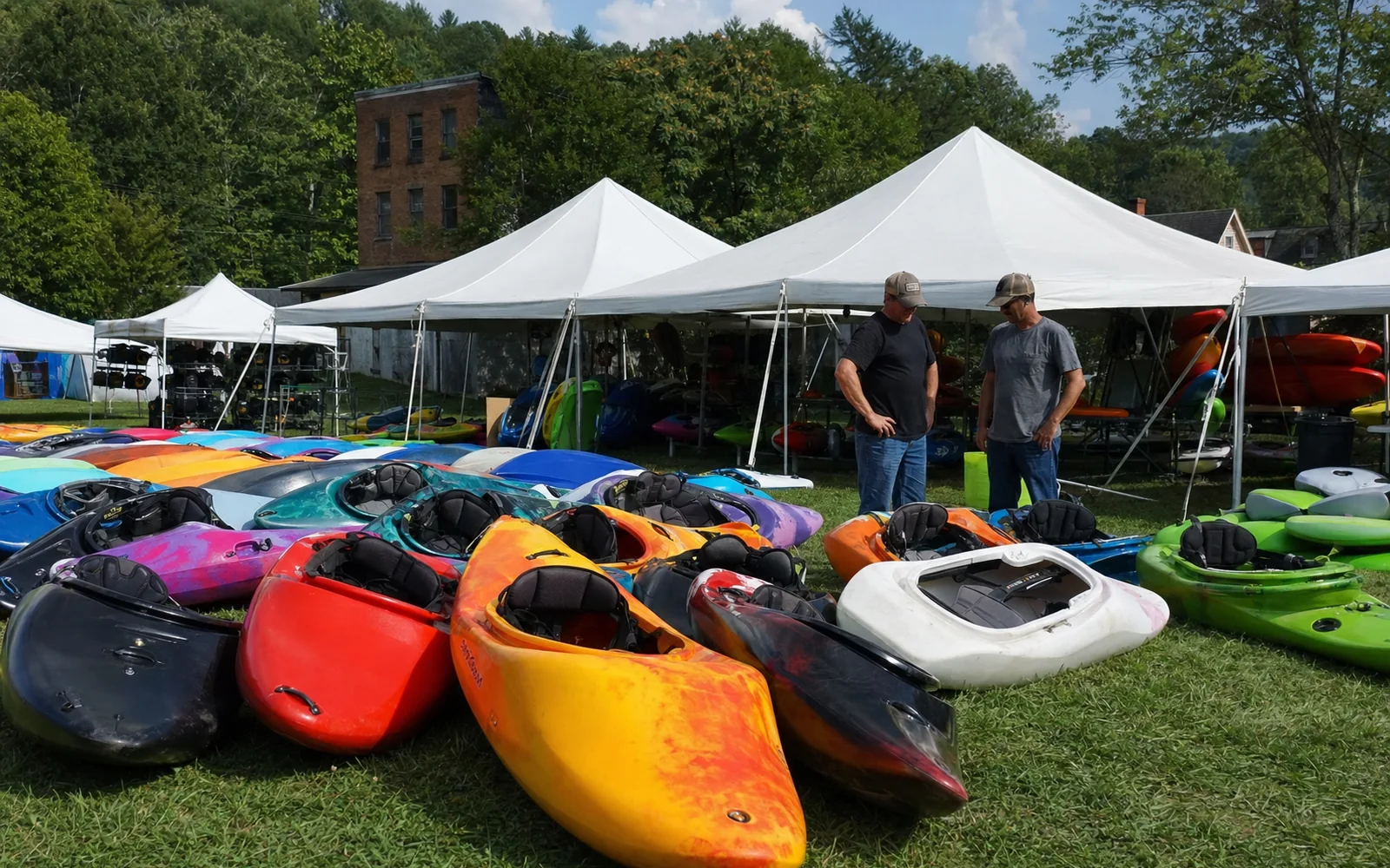 Rows of whitewater kayaks laid out on the grass at a paddling festival marketplace, white tents behind, two adults browsing, AI-generated illustration