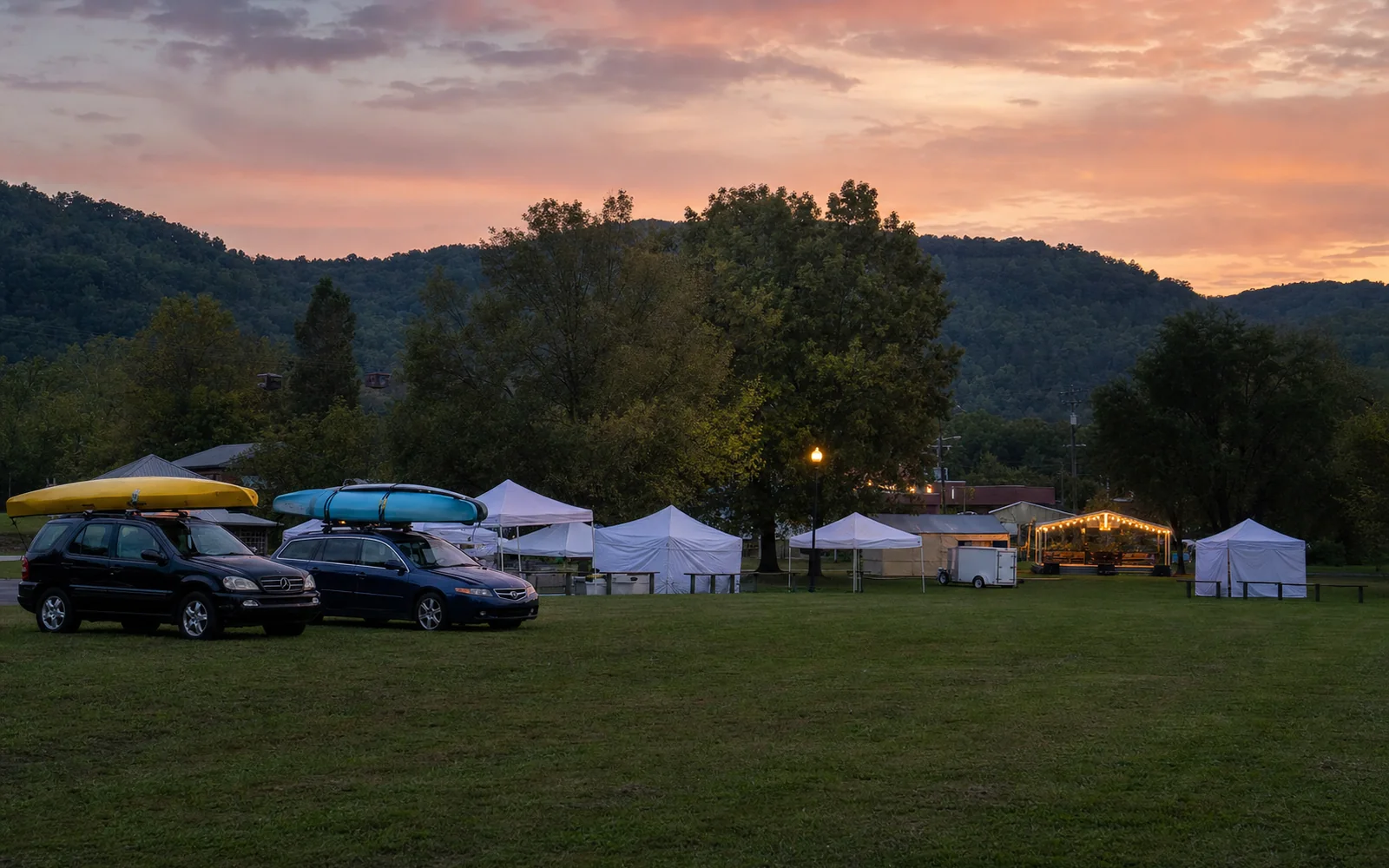 A grass festival field at dusk with white vendor tents, a small stage lit with string lights and two cars carrying whitewater kayaks on their roofs, wooded hills behind, AI-generated illustration