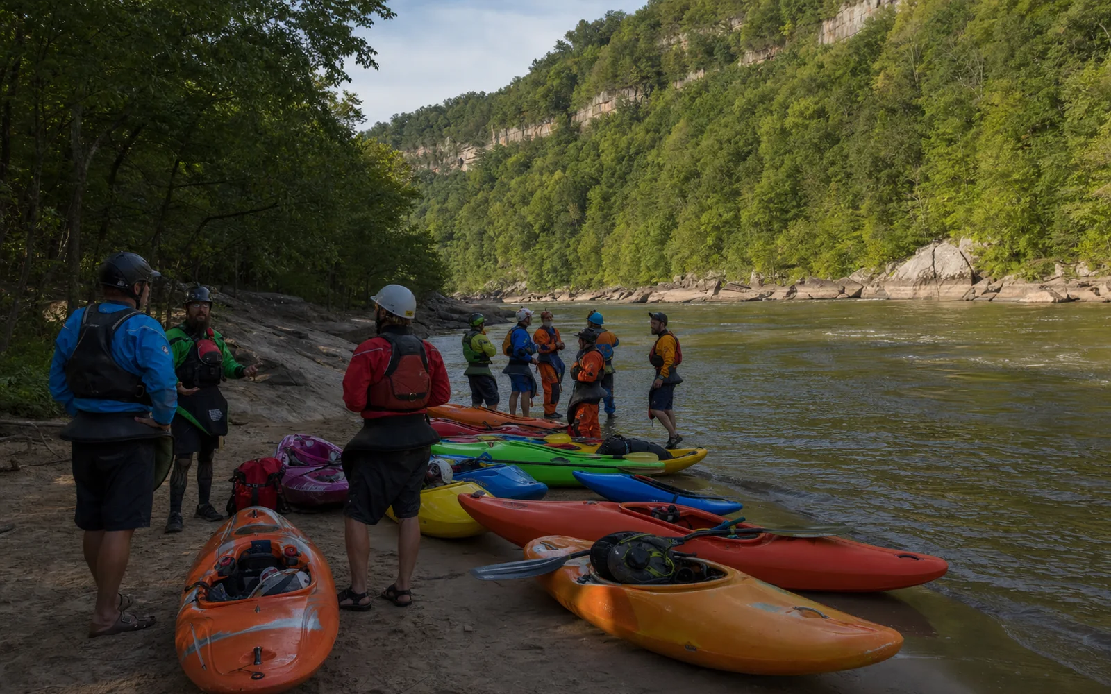 Adult paddlers in helmets and drytops talking beside a row of colorful whitewater kayaks pulled up on a sandy river beach under the forested cliffs of the gorge, AI-generated illustration