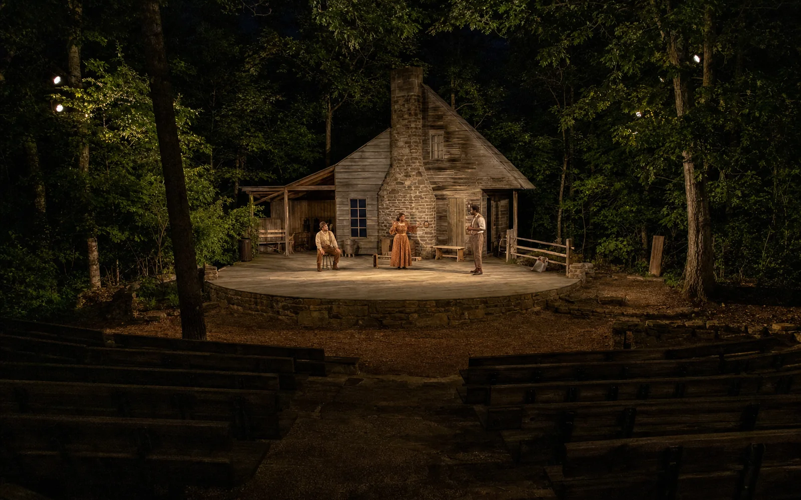 An outdoor amphitheatre stage in the forest lit at night, with three actors in 19th-century Appalachian costume in front of a log cabin set and empty benches in the foreground, AI-generated illustration