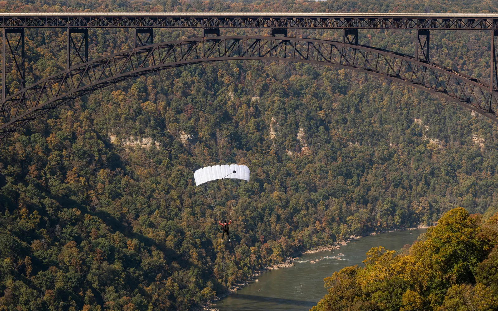 A single BASE jumper under a white canopy drifting far below the steel truss arch of the New River Gorge Bridge, the river and autumn forest below, AI-generated illustration