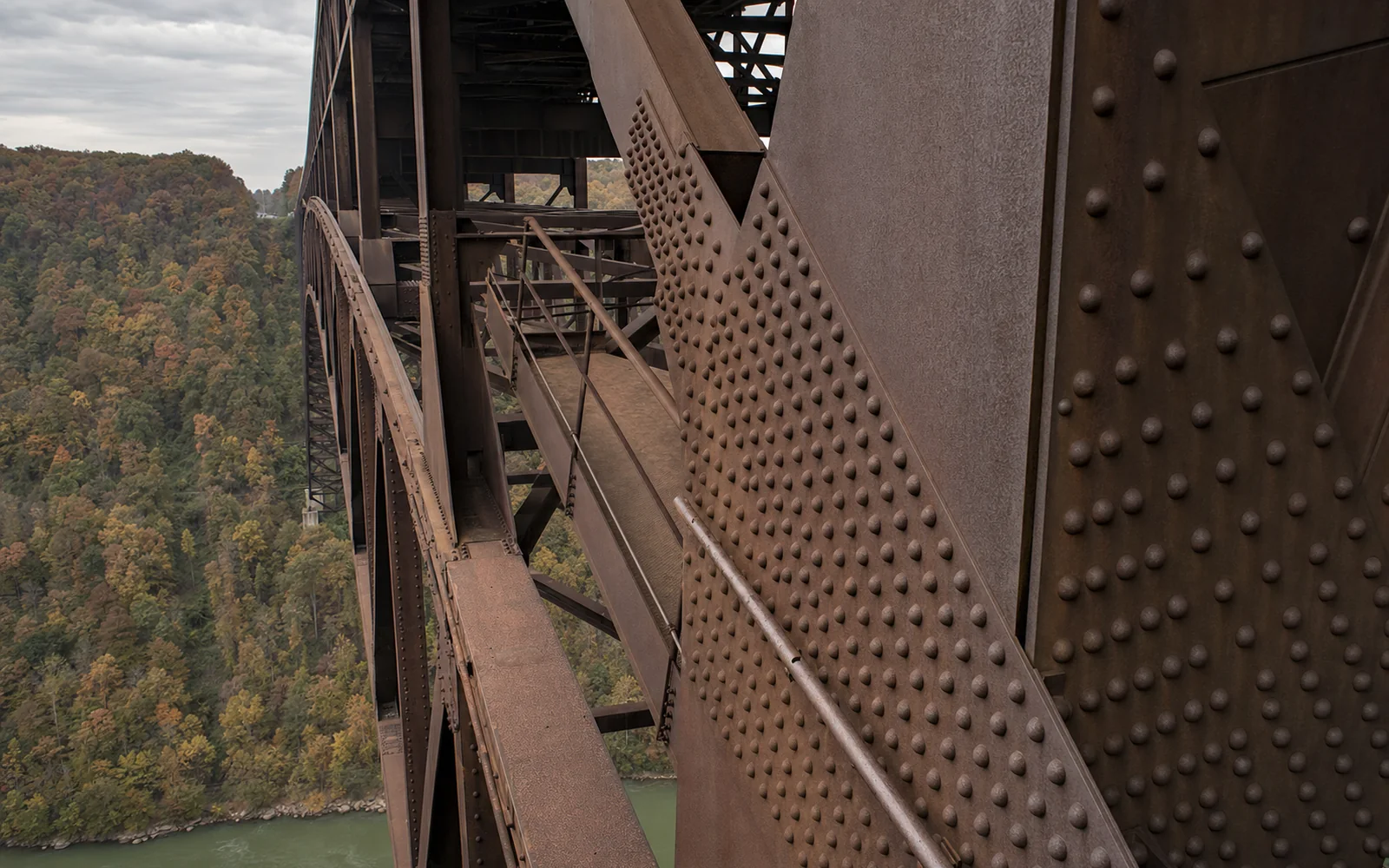 Close view of the New River Gorge Bridge's arch steel from catwalk level, riveted plates weathered brown, the deck overhead and the forested gorge falling away below, AI-generated illustration
