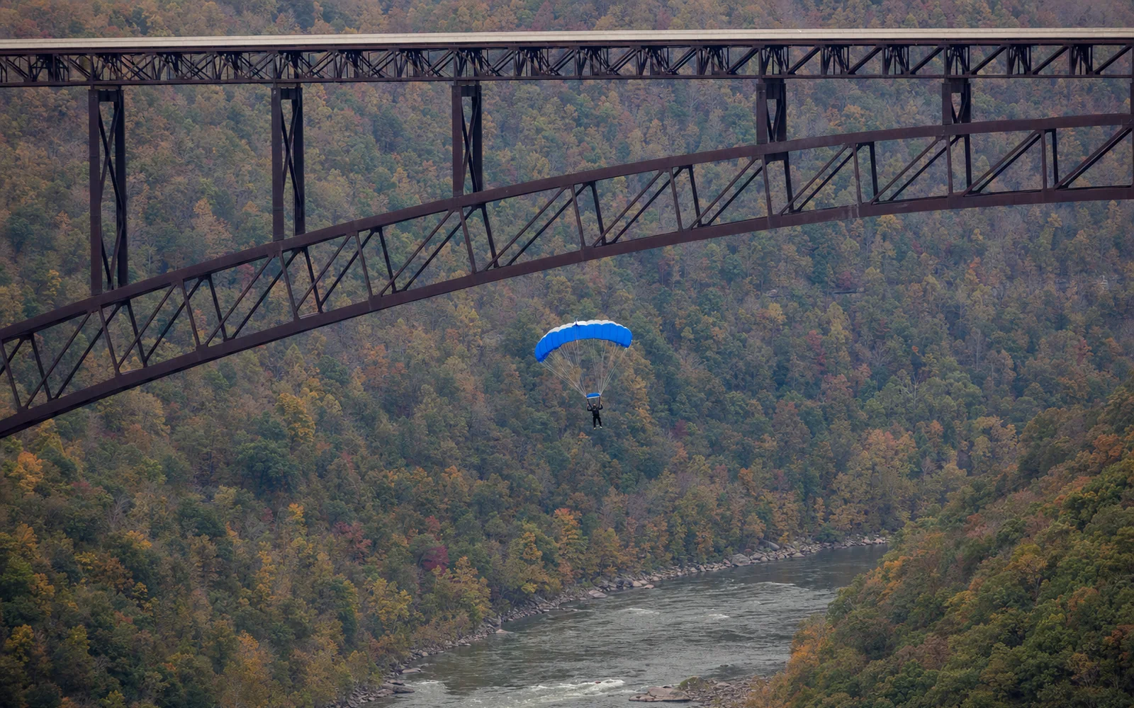 A BASE jumper under a blue canopy drifts toward the river below the steel arch of the New River Gorge Bridge, with early fall color on the forested walls of the gorge, AI-generated illustration