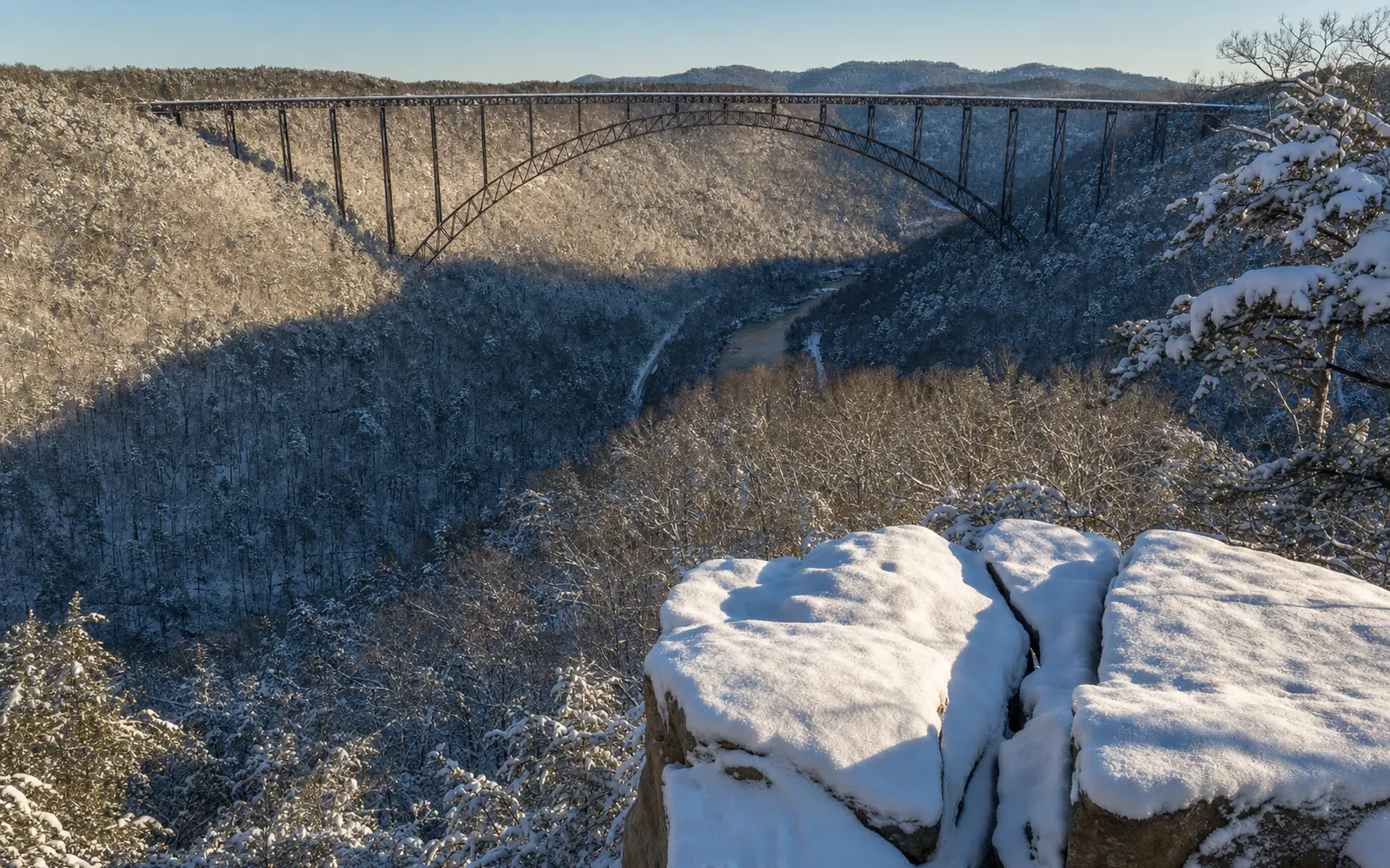 Snow on the rim at the Long Point overlook, the bare sandstone point dusted with snow, snow-laden hemlocks and bare oaks, the rust-brown steel arch of the New River Gorge Bridge straight ahead across the gorge, AI-generated illustration