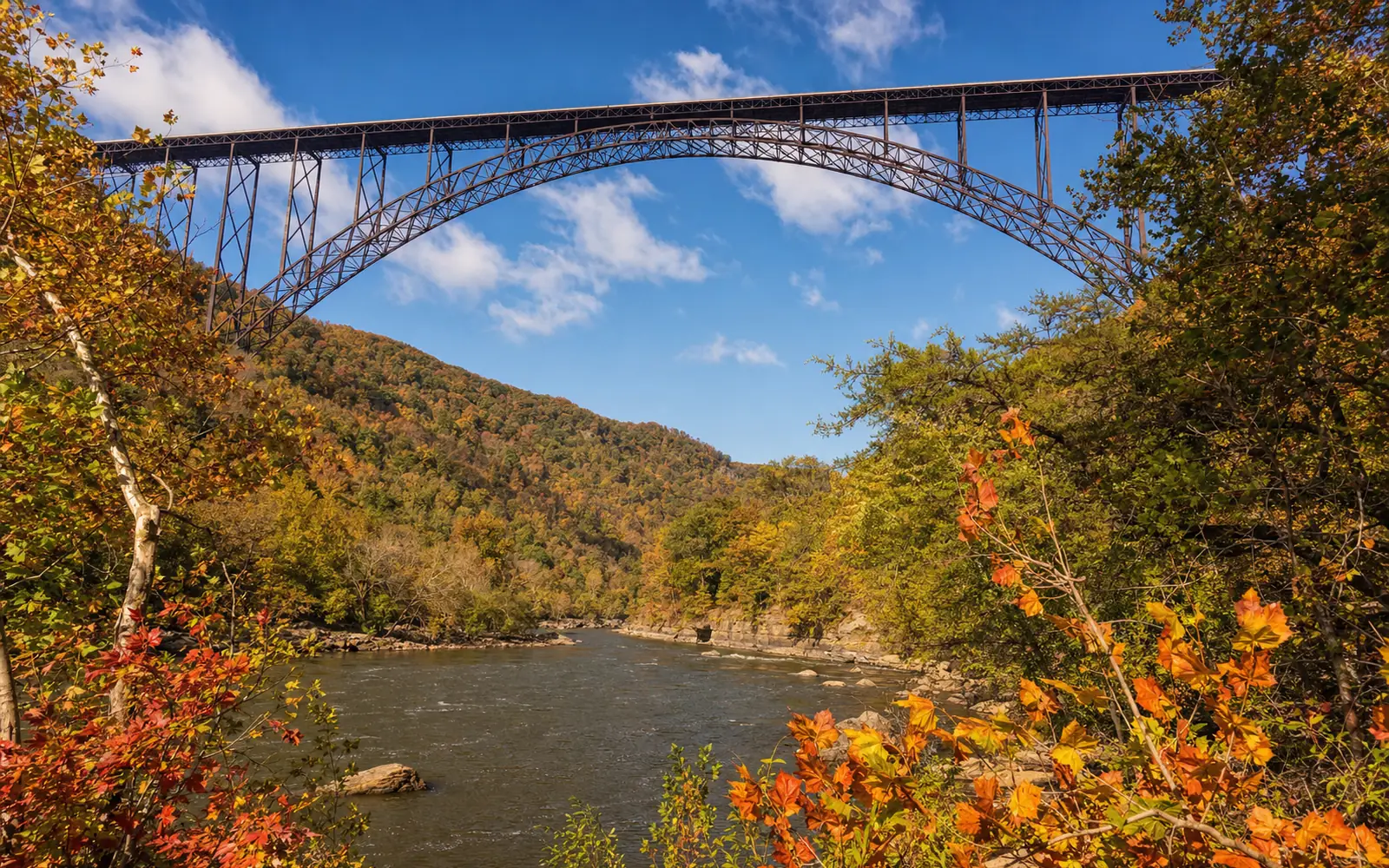 Looking up at the New River Gorge Bridge's arch from the river bank at Fayette Station through red and gold autumn foliage, AI-generated illustration