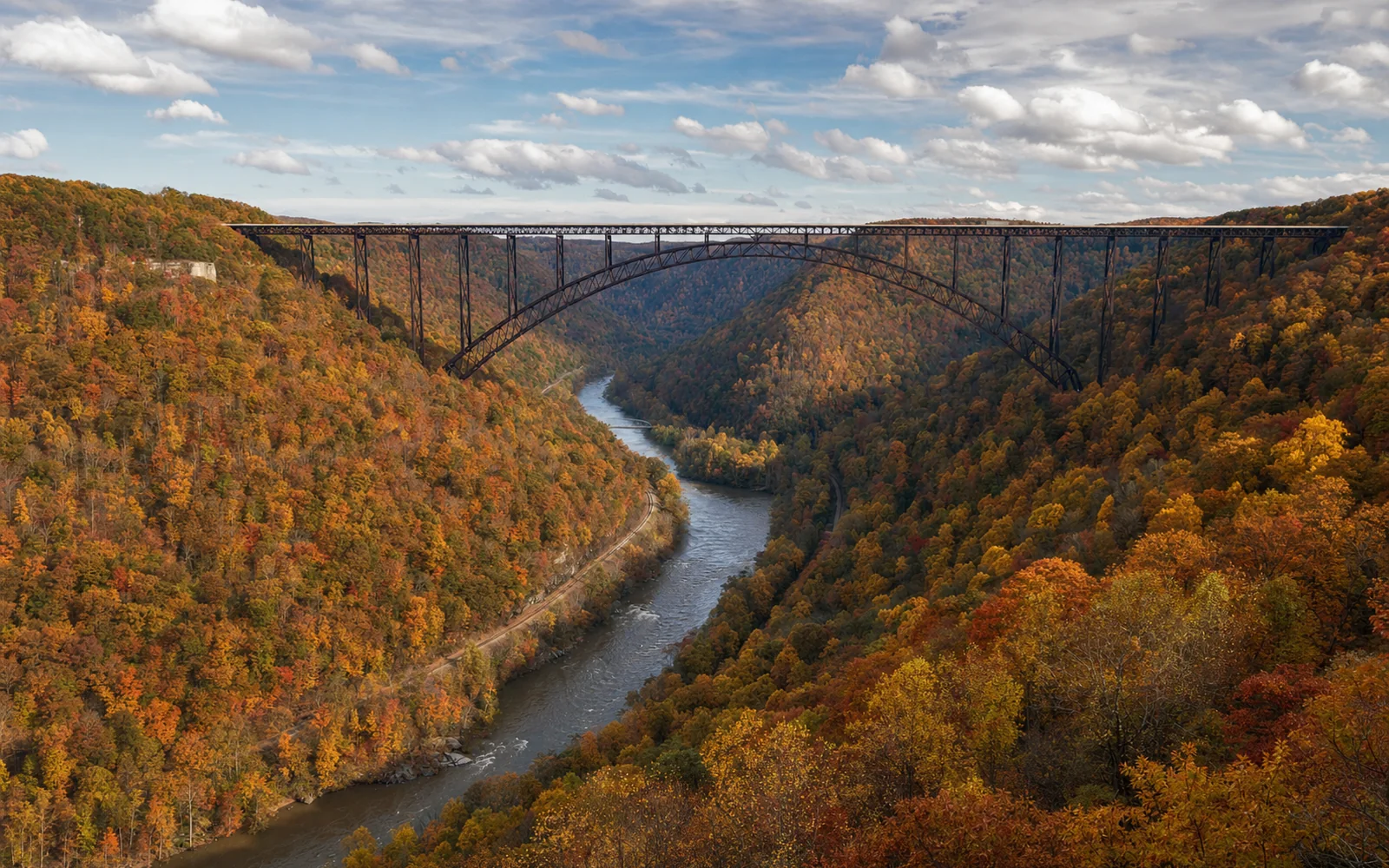 The rust-brown arch of the New River Gorge Bridge crossing the gorge in peak autumn color, red and gold slopes seen from the Canyon Rim overlook, AI-generated illustration