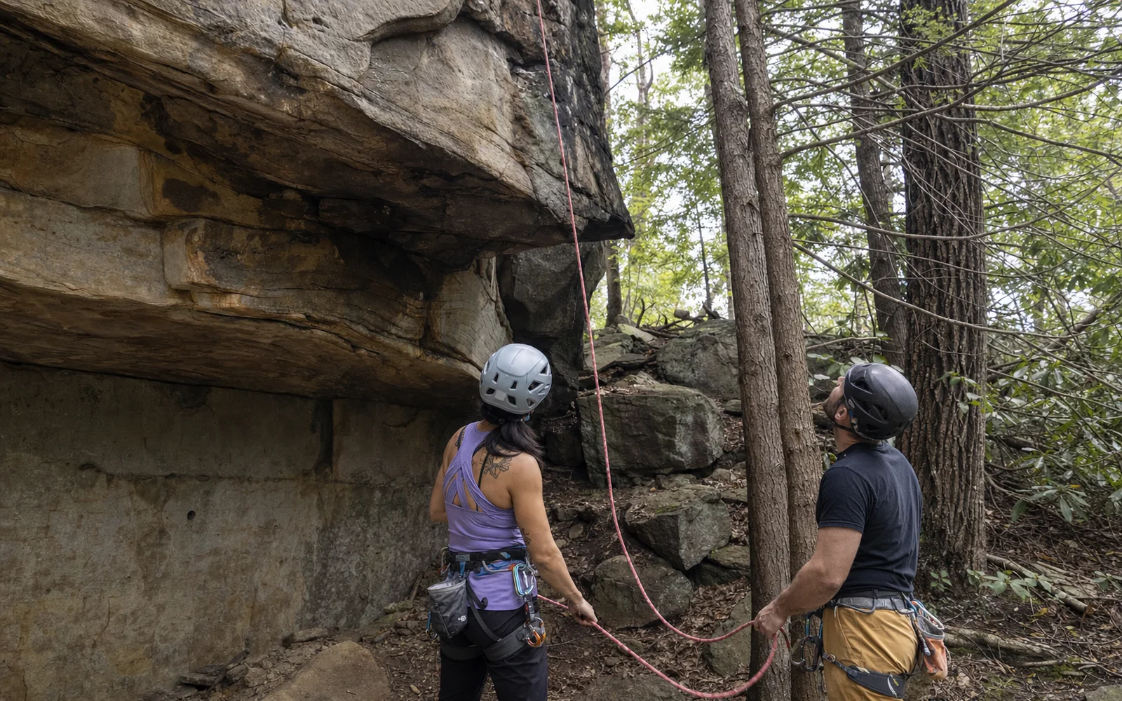 Two adults at the base of a sandstone cliff flaking a rope, helmets on, the cliff rising above, AI-generated illustration
