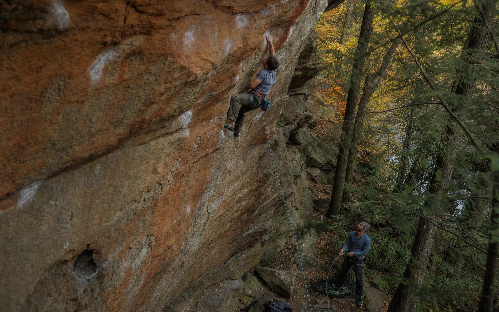 An adult climber on an overhanging orange and gray sandstone wall with chalked holds, roped to an adult belayer standing below among hemlocks and early autumn trees, AI-generated illustration