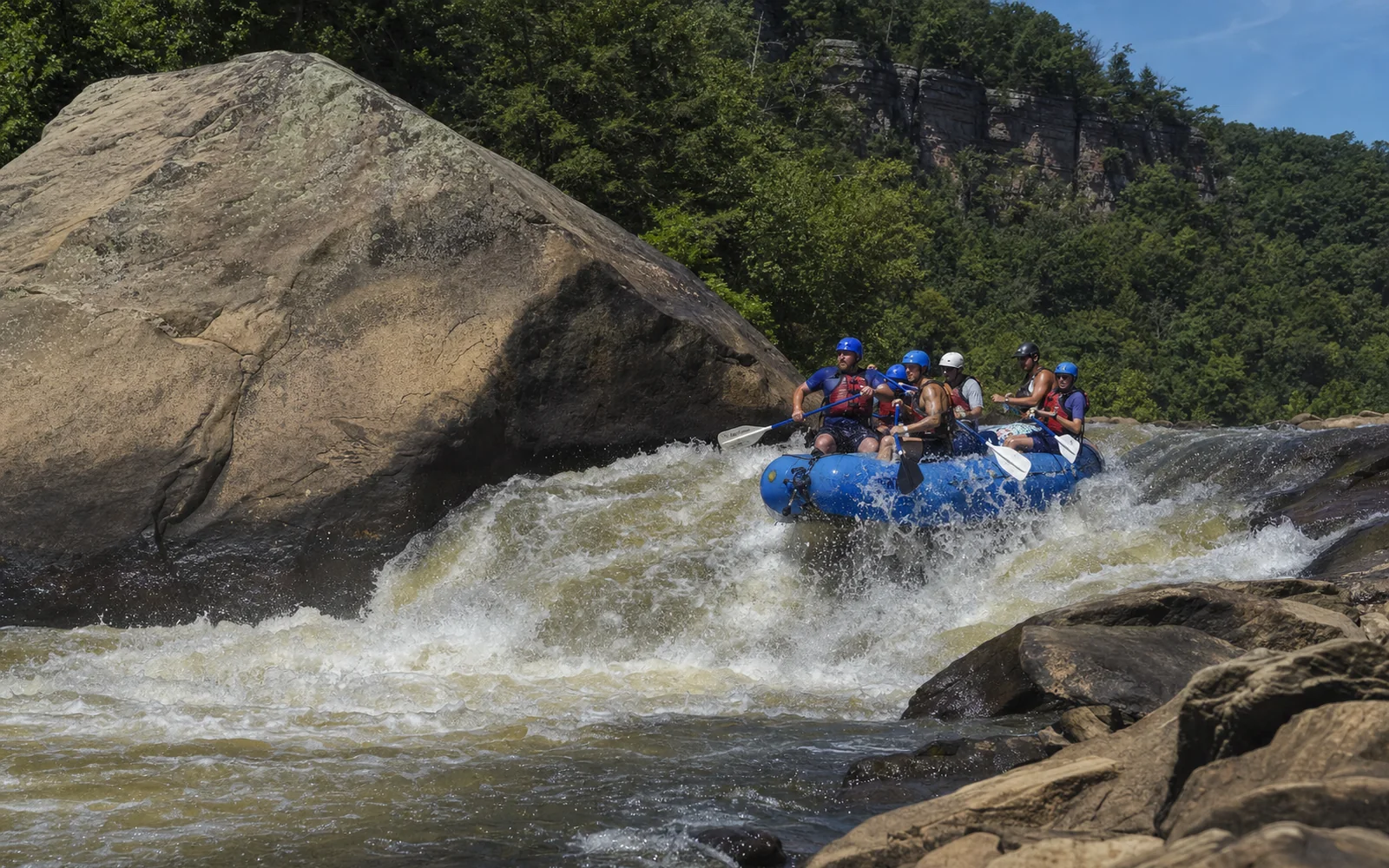 A blue commercial raft of adult paddlers in helmets and life jackets dropping through a big brown-green rapid on the Lower New River, a sandstone boulder the size of a house at left, AI-generated illustration