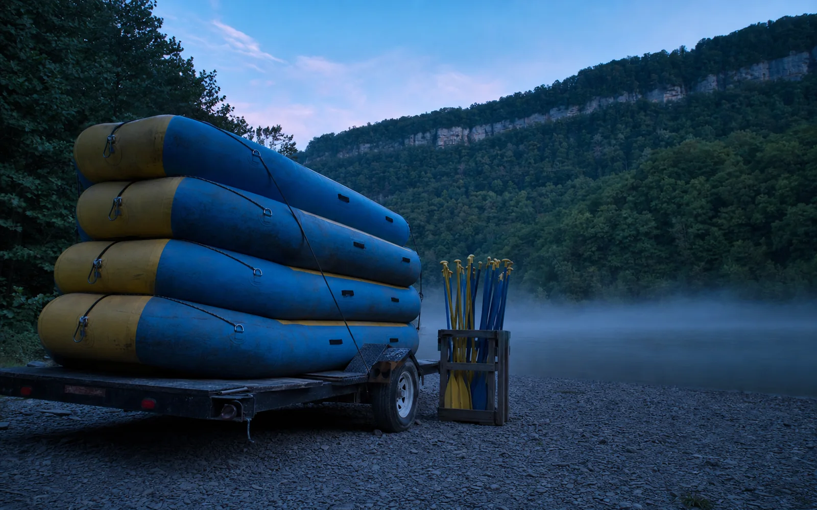 Blue and yellow whitewater rafts stacked upside down on a flatbed trailer at a gravel river put-in at dawn, a crate of paddles beside it and mist on the river, AI-generated illustration