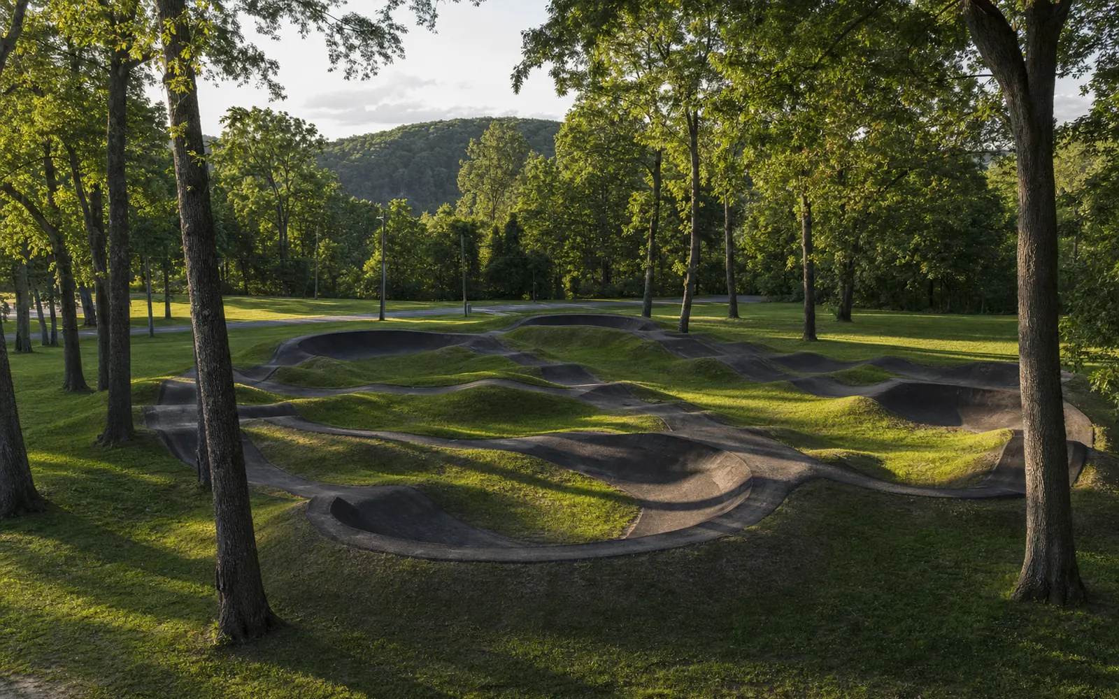 A paved bicycle pump track with rolling berms and bumps in a mown town park among oak trees, AI-generated illustration