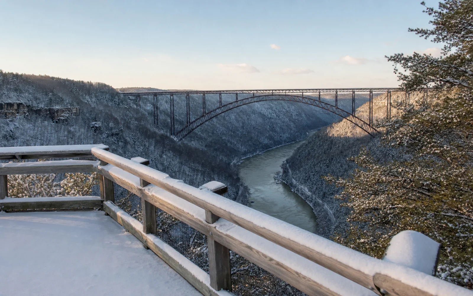 The Canyon Rim upper overlook deck in winter, snow on the timber railings and deck, the New River Gorge Bridge's steel arch dark against a pale sky, frosted forest, AI-generated illustration