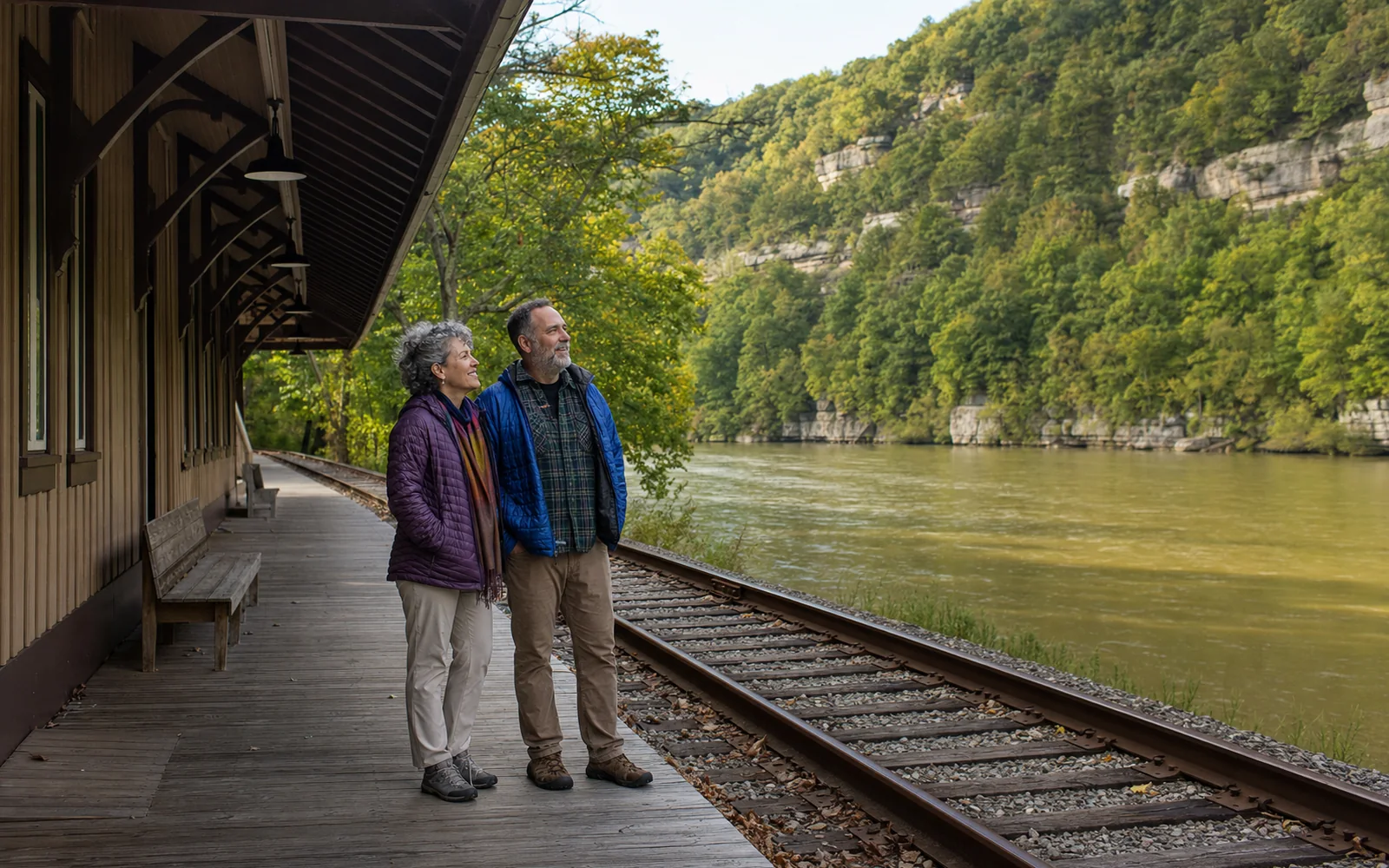 An adult couple in their fifties standing on the platform of the restored Thurmond depot, the railroad tracks and the river beside them, AI-generated illustration