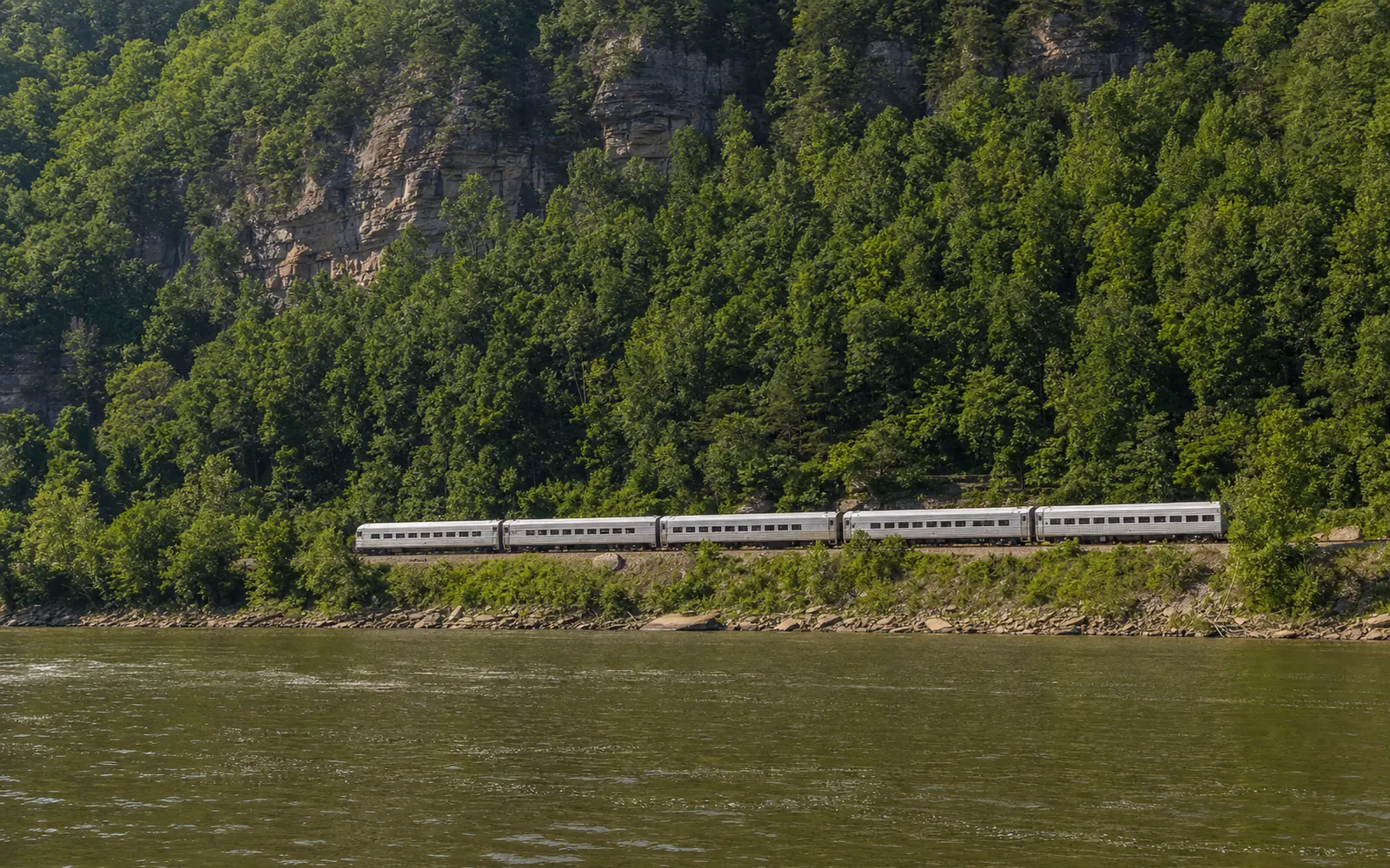 A silver passenger train curving along the New River at the bottom of the gorge on a narrow ledge between the river and a wooded slope, AI-generated illustration