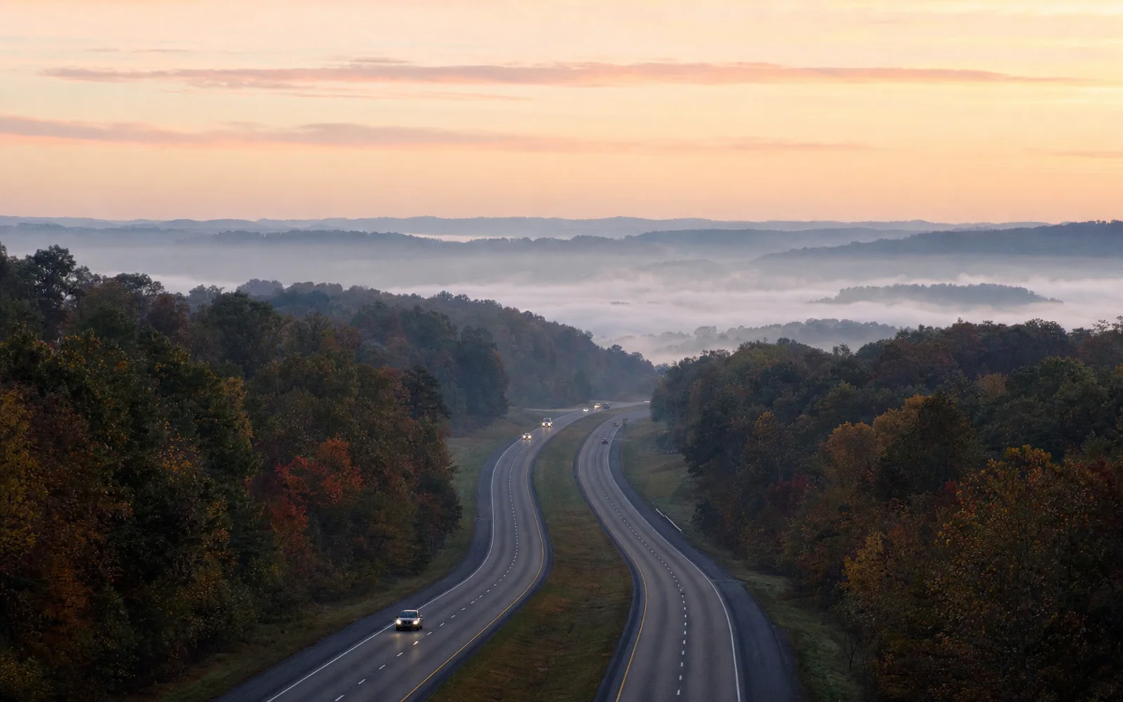 The divided four-lane U.S. 19 at dawn sweeping over wooded plateau hills, fog lying in the hollows, sparse traffic with headlights, AI-generated illustration