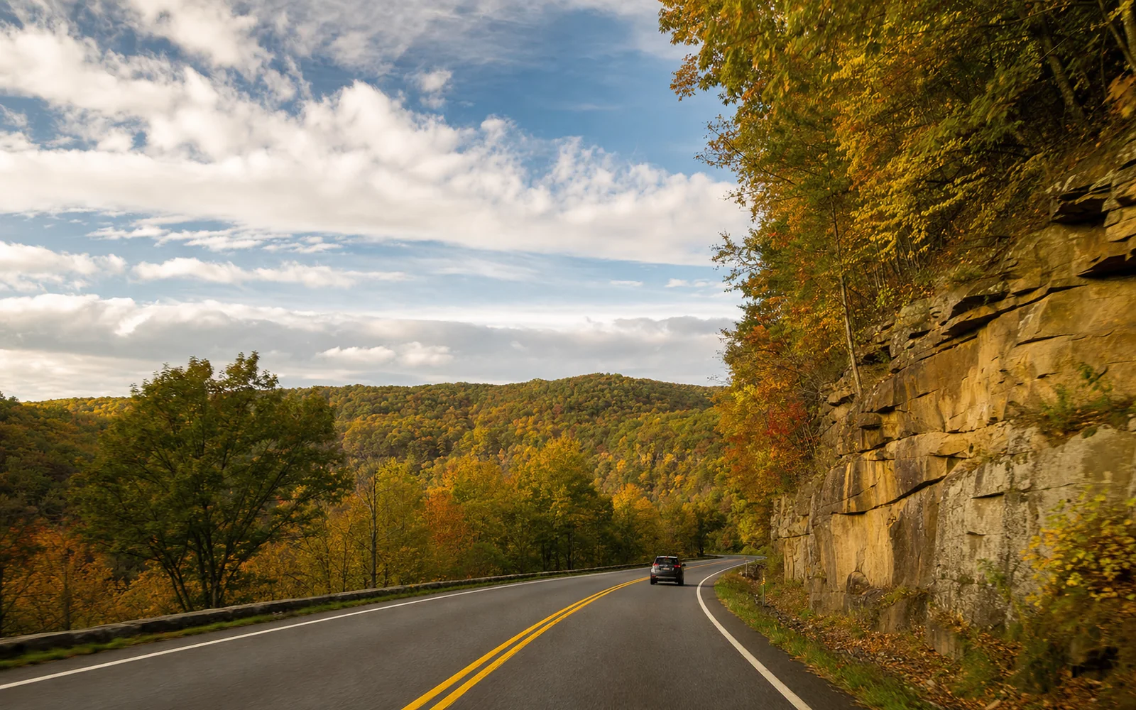 A two-lane mountain highway curving past a layered sandstone road cut and hardwood ridges in yellow and orange fall color, with one car ahead under a partly cloudy sky, AI-generated illustration