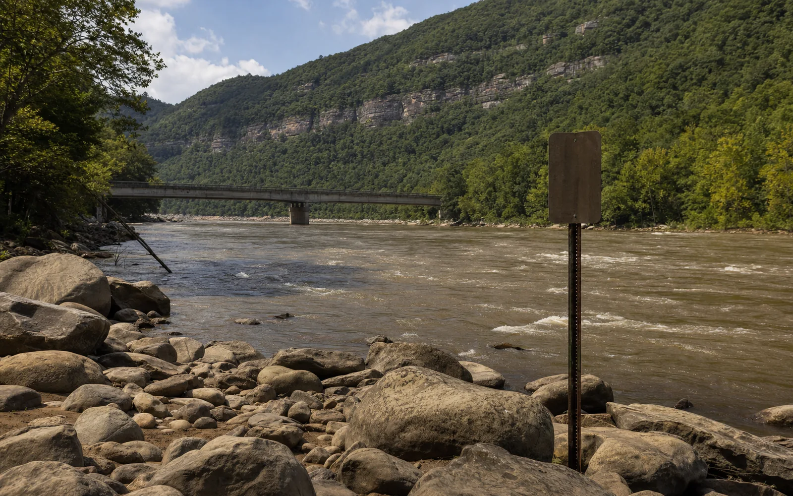 A rocky riverbank of rounded boulders beside a fast brown river with small rapids, a blank sign on a metal post, and a low concrete bridge crossing below a forested gorge wall, AI-generated illustration