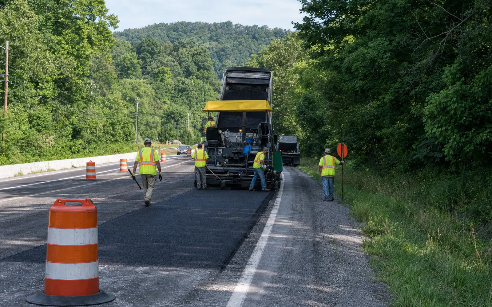 A paving crew of adults in high-visibility vests working one lane of a four-lane mountain highway, orange barrels, a flagger, wooded hills, AI-generated illustration