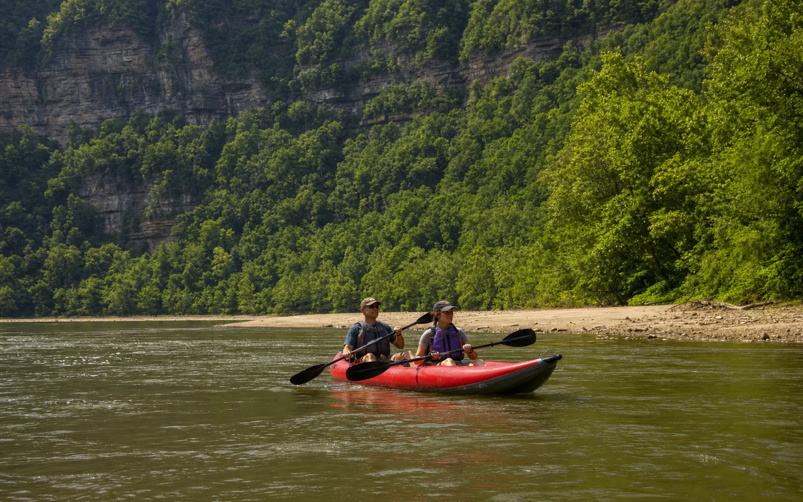 Two adults in life jackets paddling a red inflatable kayak on calm brown water below a forested sandstone cliff, a sandy bank behind them, AI-generated illustration