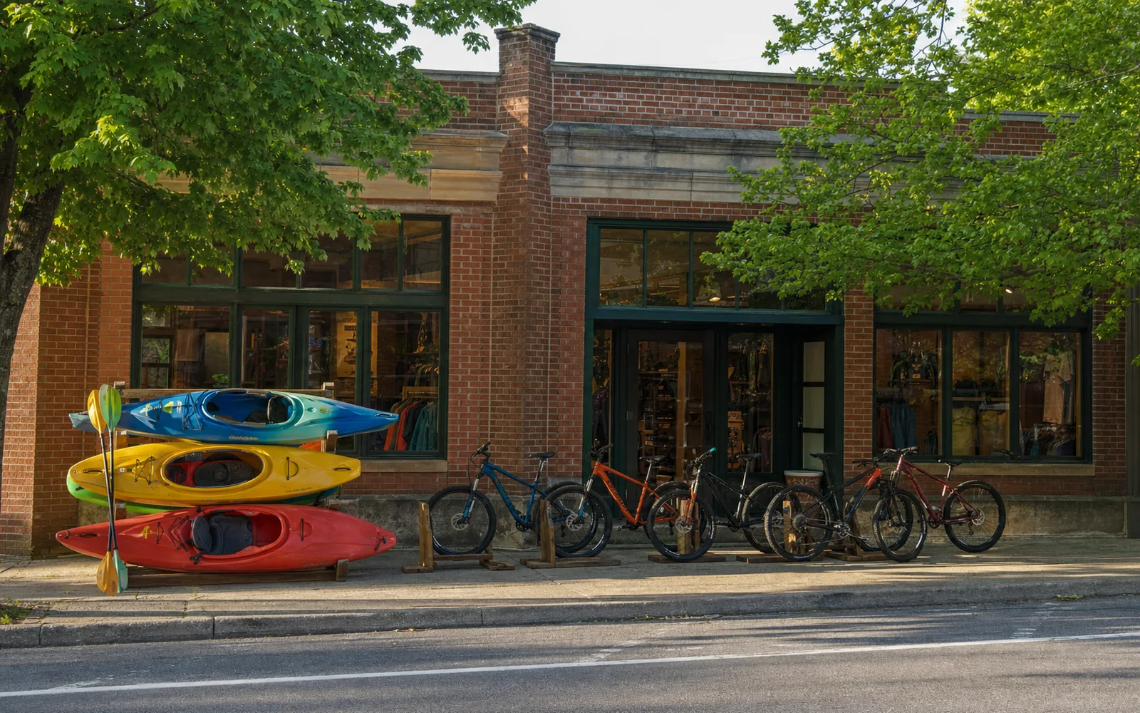 A brick storefront on a tree-lined downtown street with whitewater kayaks stacked on a rack and mountain bikes lined up on the sidewalk outside, no people, AI-generated illustration