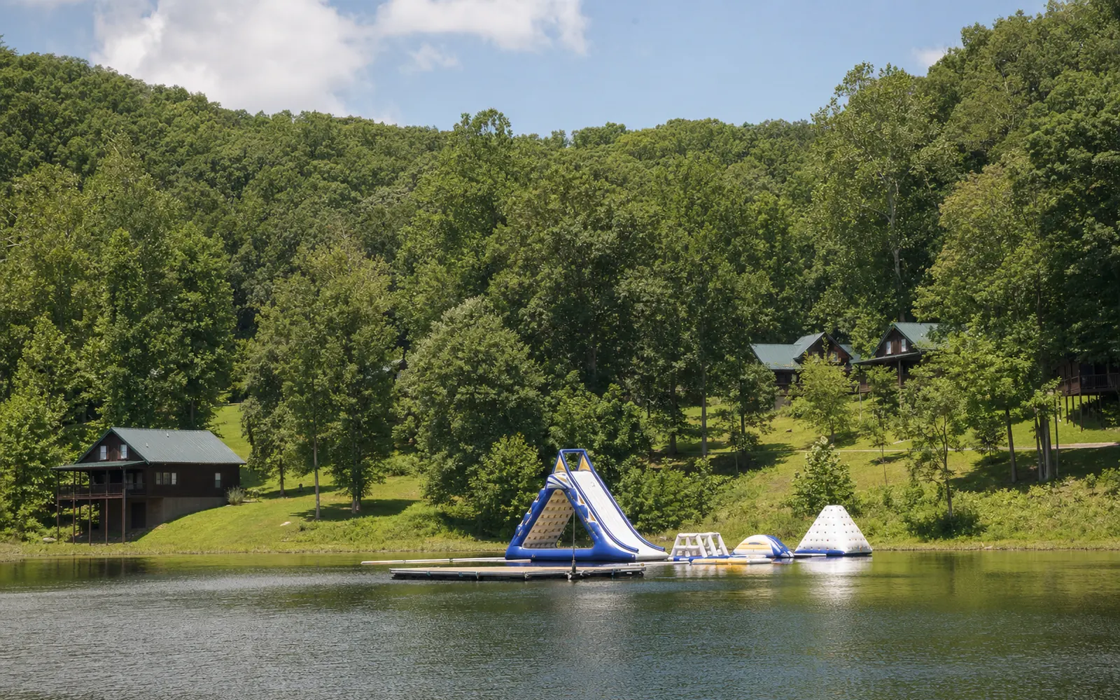 A small resort lake at dawn with a floating platform and inflatable water slides on calm water, cabins among oak and hickory trees behind, no people, AI-generated illustration