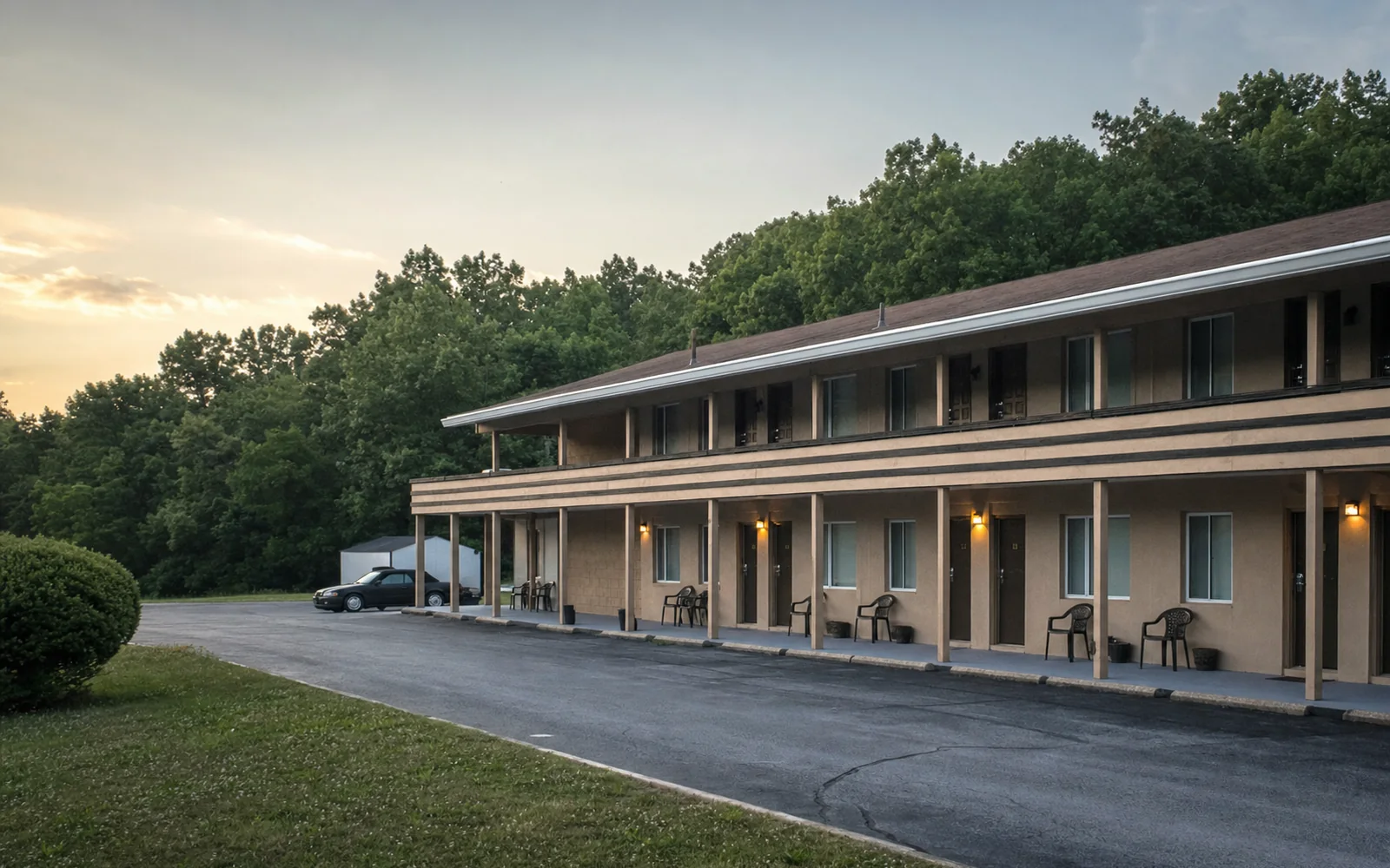 A two-story motor inn with a covered walkway and chairs outside the rooms, a paved lot and wooded hills behind at dusk, AI-generated illustration