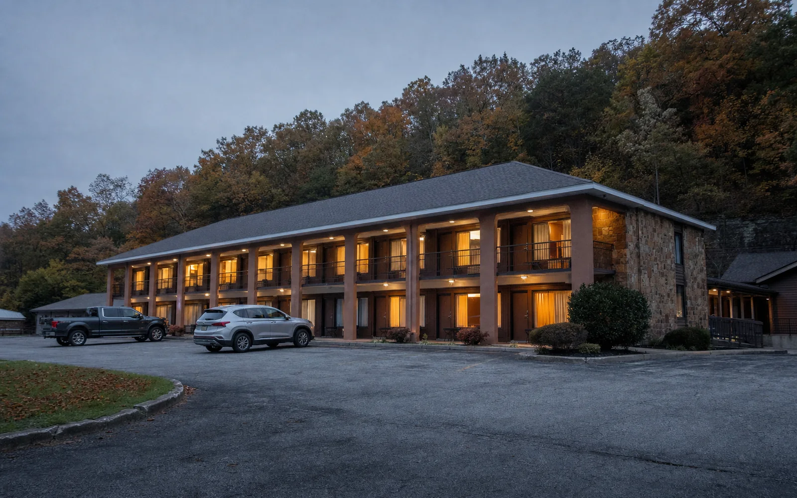 A two-story highway hotel at dusk with lit windows and a small parking lot, a forested ridge behind it, AI-generated illustration